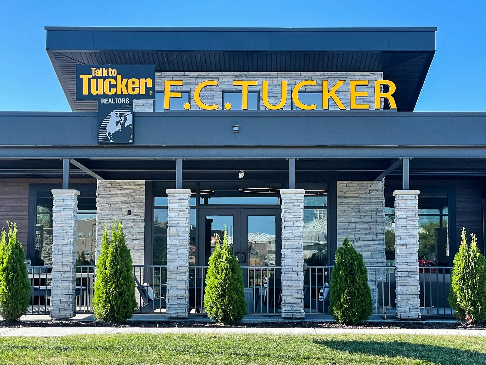 Exterior view of a modern real estate office with the sign 'F.C. Tucker Realtors' and a smaller sign 'Talk to Tucker' with a logo of a globe and the words 'REALTORS'. There are five stone columns, green bushes, and a glass entrance door among the bui