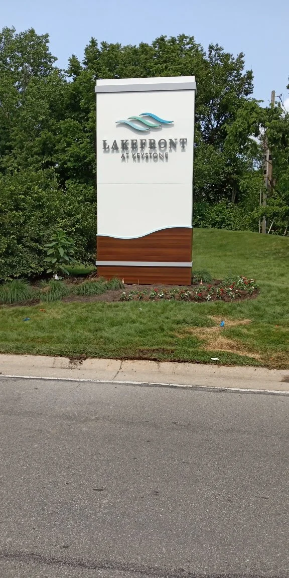 Sign for Lakefront at Keystone community, featuring a logo with blue waves and black text, surrounded by greenery and a landscaped area.