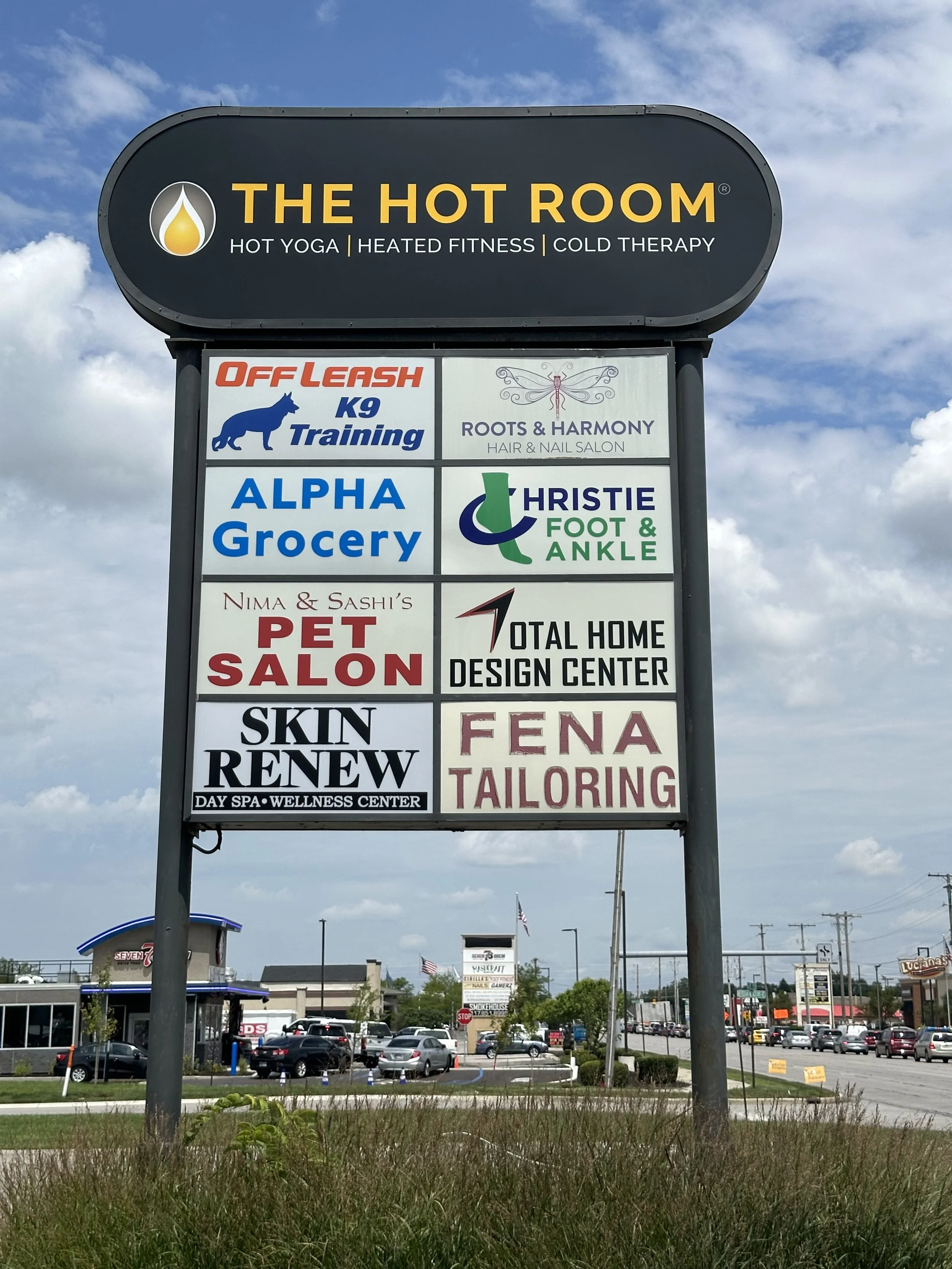 A large outdoor signboard displaying multiple business advertisements, including THE HOT ROOM for hot yoga, heated fitness, and cold therapy, along with other businesses such as dog training, flower and hair salon, grocery store, foot and ankle clini