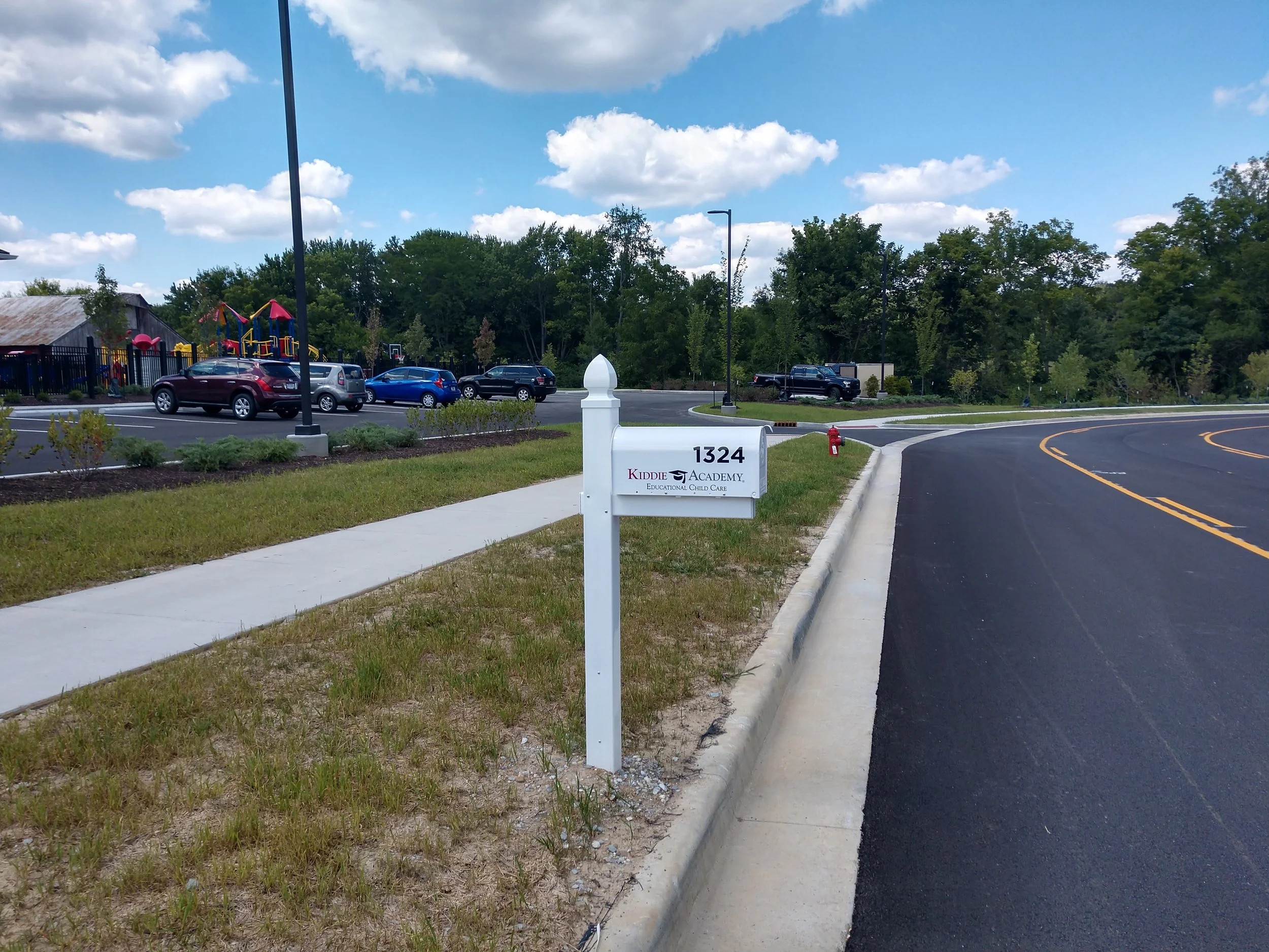A white mailbox with the address 1324 and a sign for Kiddie Academy on a grassy patch beside a sidewalk. In the background, there is a parking lot with cars, a playground with colorful equipment, and lush green trees under a partly cloudy sky.