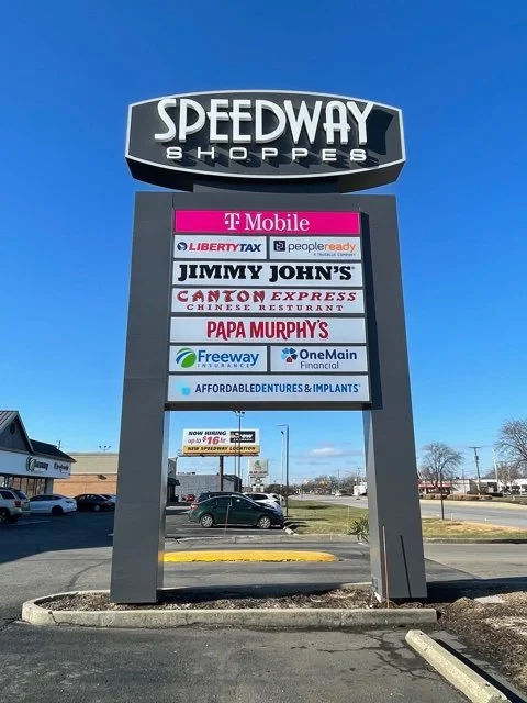 Sign for Speedway Shoppes listing various retail and restaurant businesses against a clear blue sky.