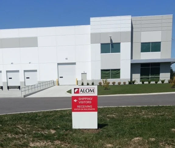 Sign for ALOM indicating shipping, visitors, and receiving entrance on Walden Road in front of a white industrial building.