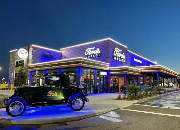 Exterior of a Ford dealership called Ford's Garage at night, with a vintage black car parked in front, illuminated by blue and white lighting.