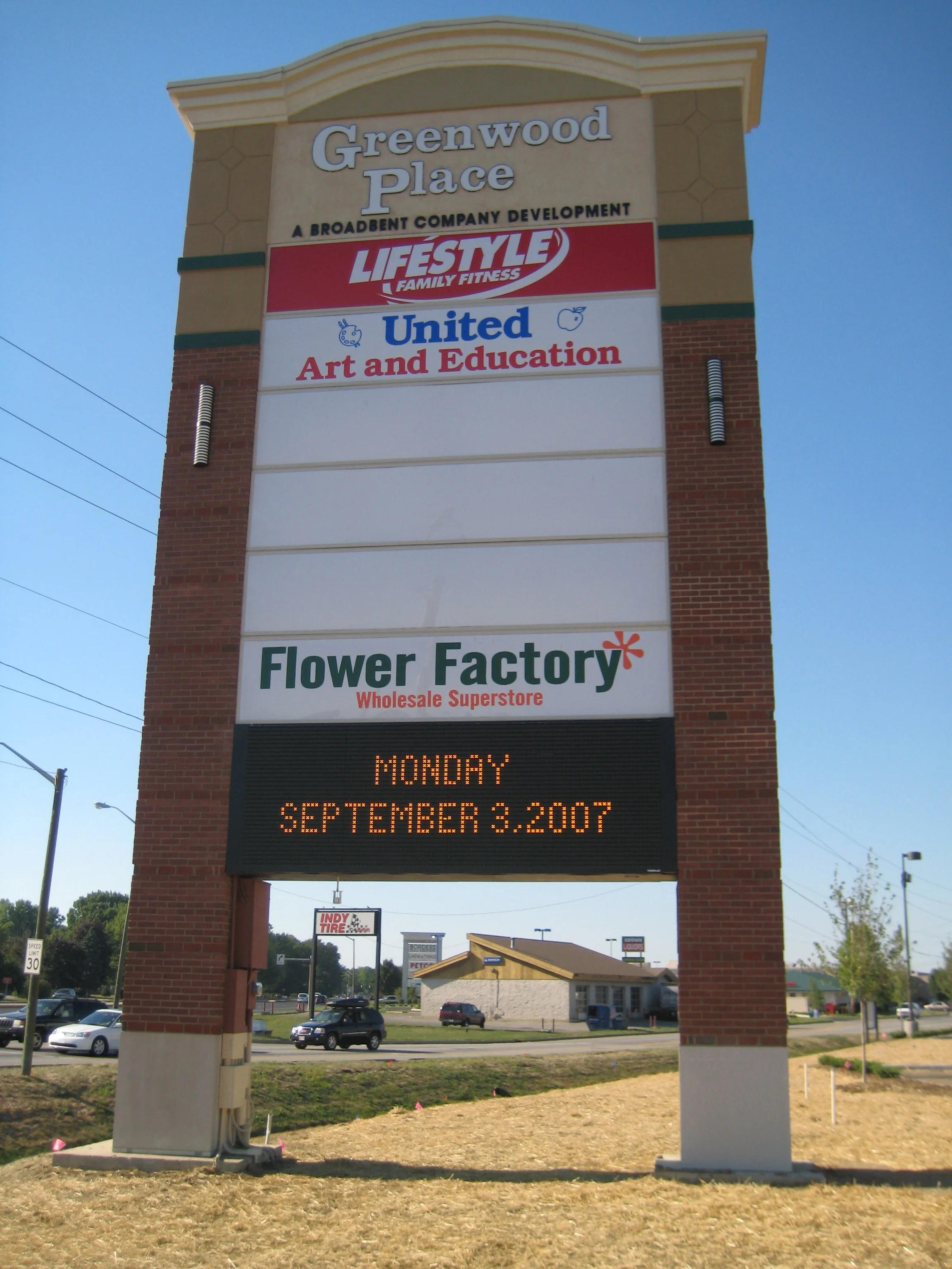 Tall billboard structure displaying multiple business signs and a digital date and day message board at the bottom. The signs include Greenwood Place, a development company; Lifestyle Family Fitness; United Art and Education; and Flower Factory Whole