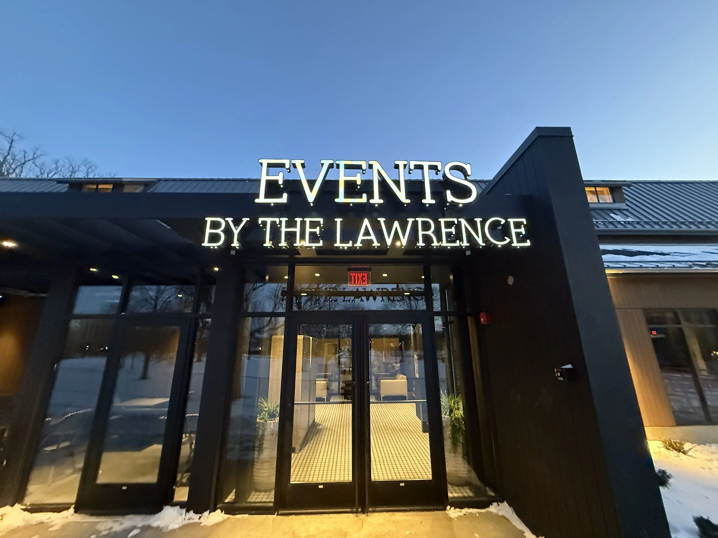 Entrance of an event venue with illuminated sign that reads 'Events by the Lawrence' during dusk, snow on the ground.