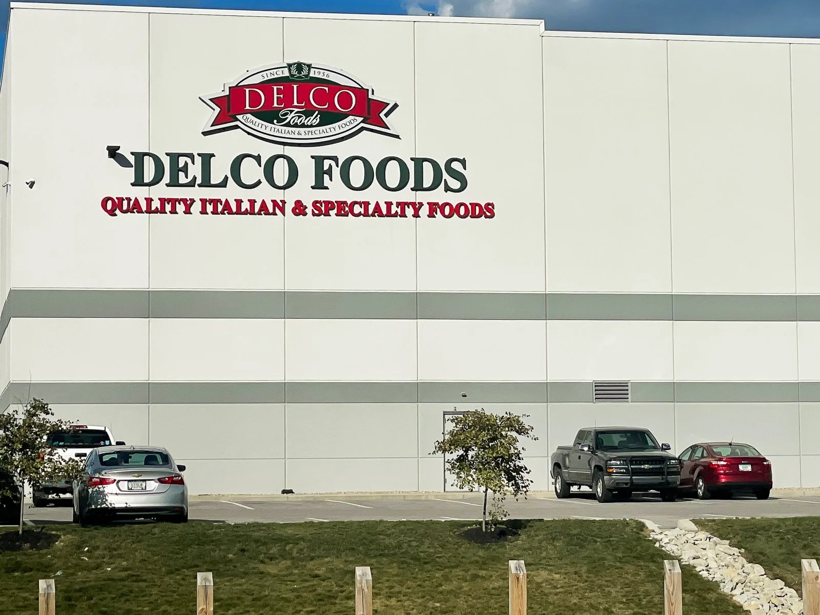 Large white building with a sign that reads "Delco Foods" and "Quality Italian & Specialty Foods." Several parked cars and small trees in front of the building.