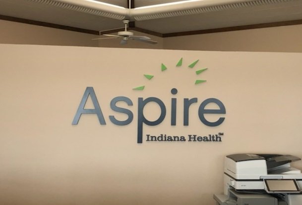 Indoor wall with the Aspire Indiana Health logo, featuring a stylized clock with green rays and blue text.