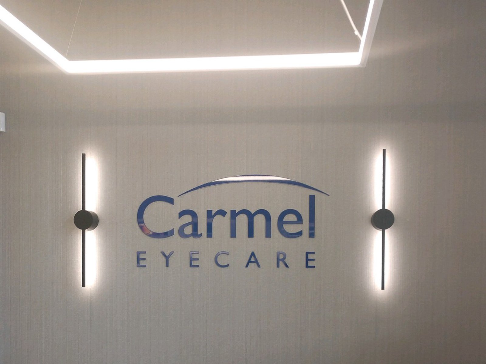 Interior wall of Carmel Eye Care with the business logo and name, modern wall-mounted lights, and a textured beige wall.