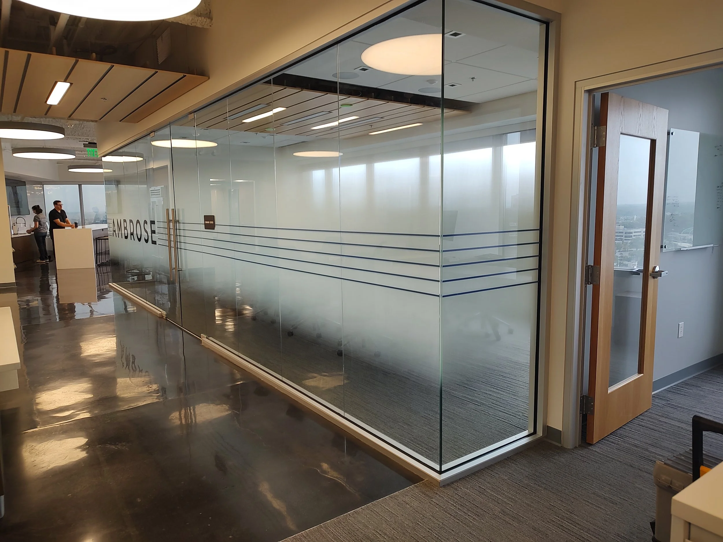 Interior office space with a glass conference room, glass walls with the word 'AMBROSE' on it, a door to the right, and people working in the background.