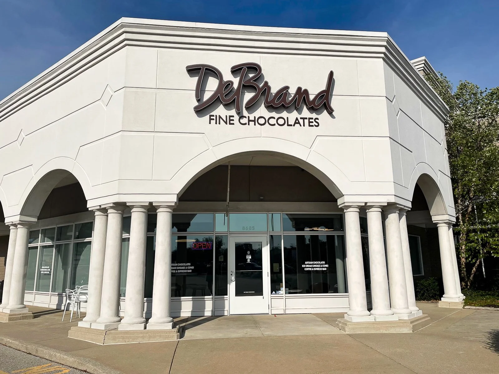 Front of a DeBrand Fine Chocolates store with white columns and large glass windows, a sign that says 'DeBrand,' and an 'Open' sign in the window.