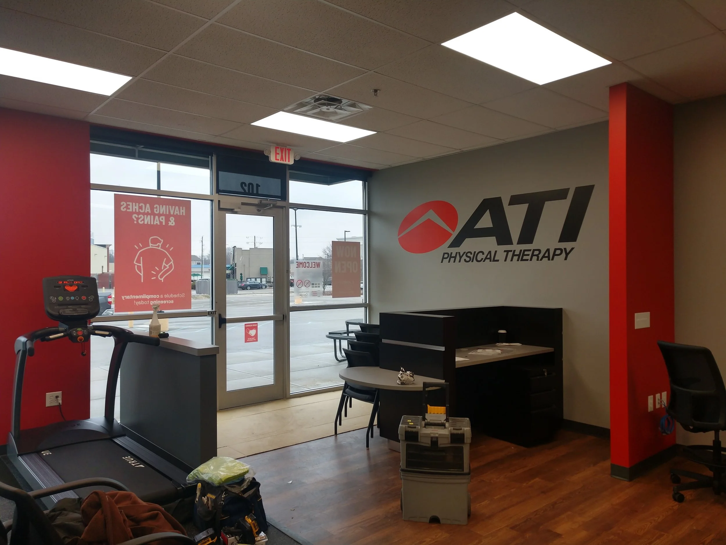 Interior of ATI Physical Therapy clinic with a treadmill, a desk, chairs, and signs on the glass doors and windows.
