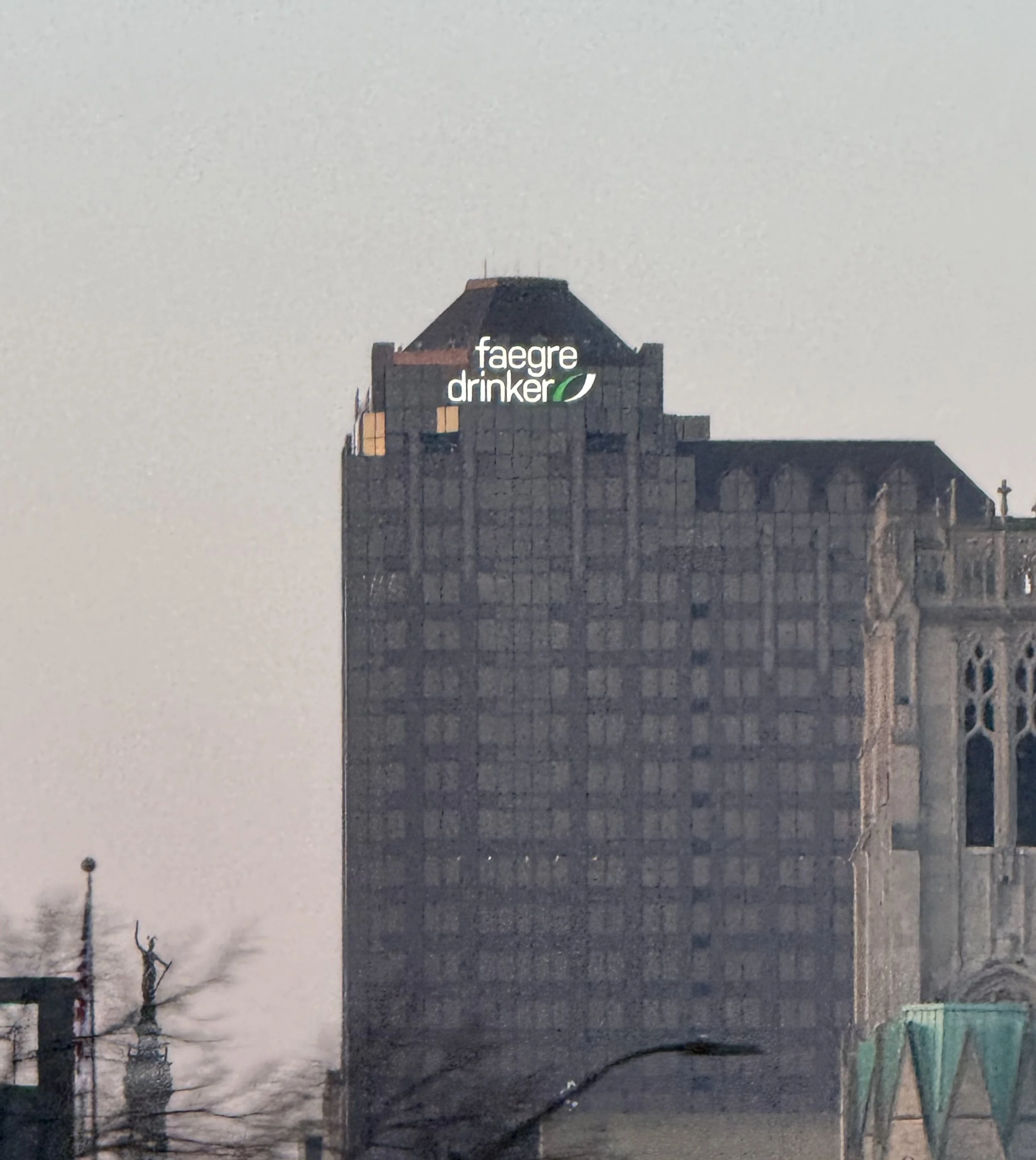 A tall building with a lit sign at the top reading 'faegre drinker' with a green leaf logo. The sky is overcast or cloudy.