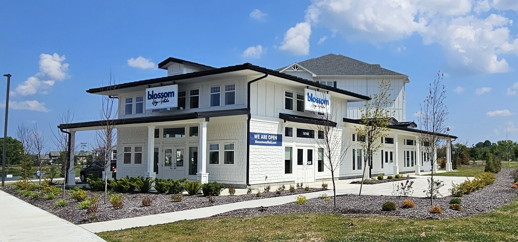 A modern, white, multi-story commercial building with signage for Blossom Your Pet, displaying a sign indicating the business is open. The building has large windows, a landscaped area with small trees and shrubs, and a sidewalk in front. Blue sky wi