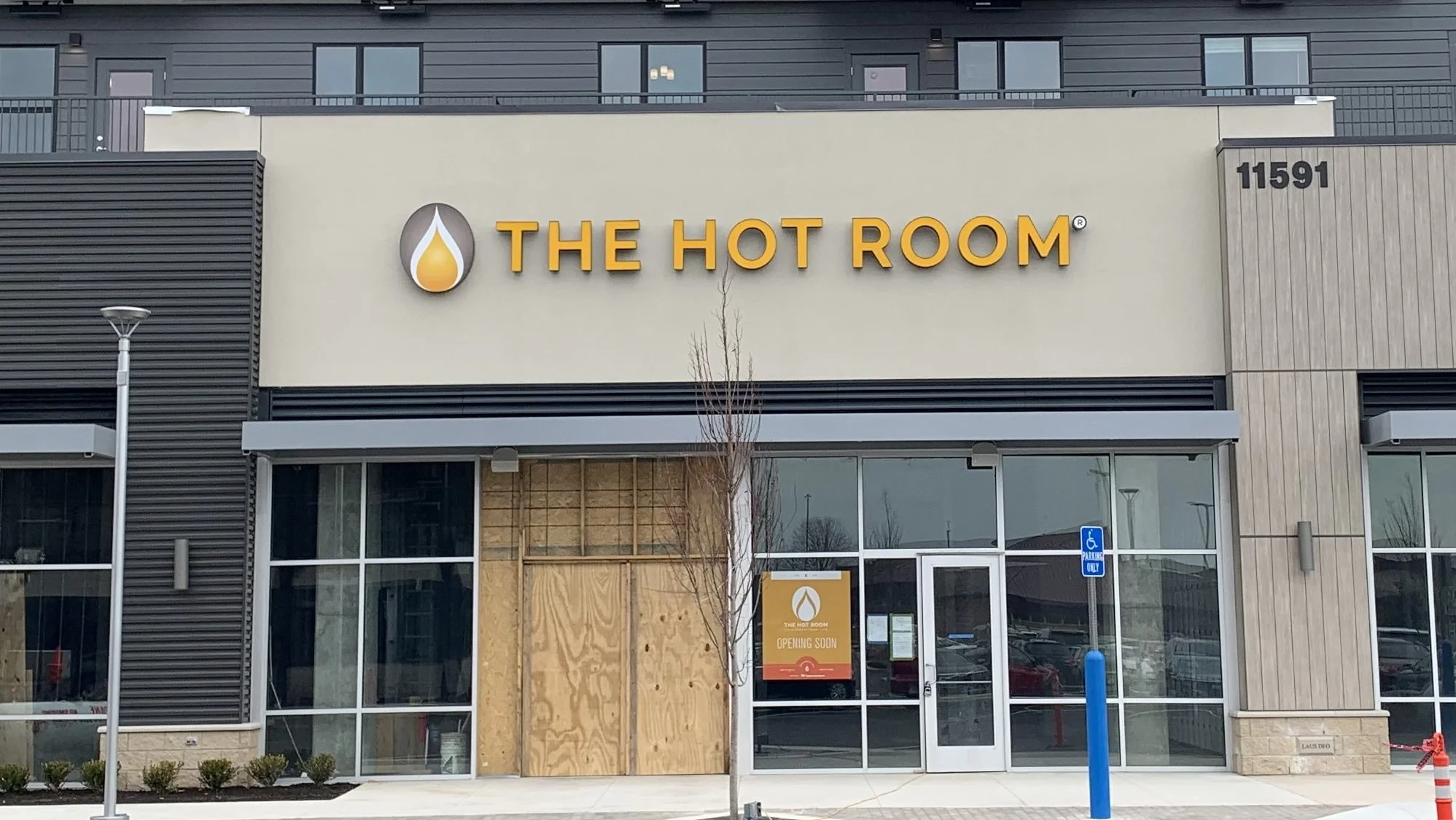 Front of a commercial building with a sign that reads "The Hot Room," with a water droplet logo next to it, and a sign indicating it is opening soon. The building has black and beige exterior walls, large glass windows, and a sidewalk in front with a