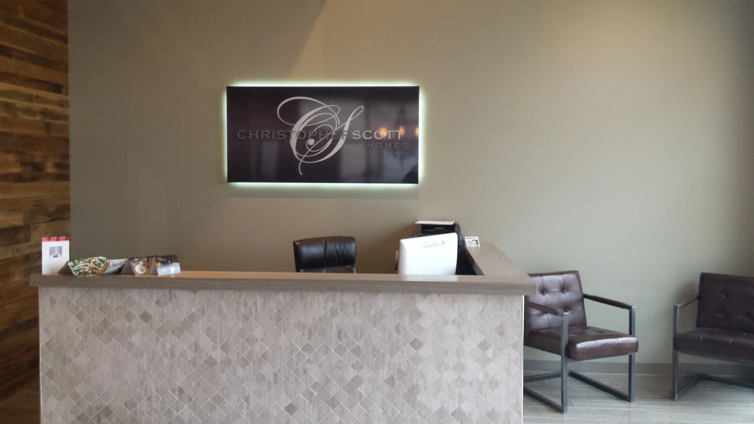 Office reception area with a beige patterned front desk, a black leather chair behind the desk, a computer, and two brown chairs with metal frames to the right, against beige and wooden wall backgrounds.