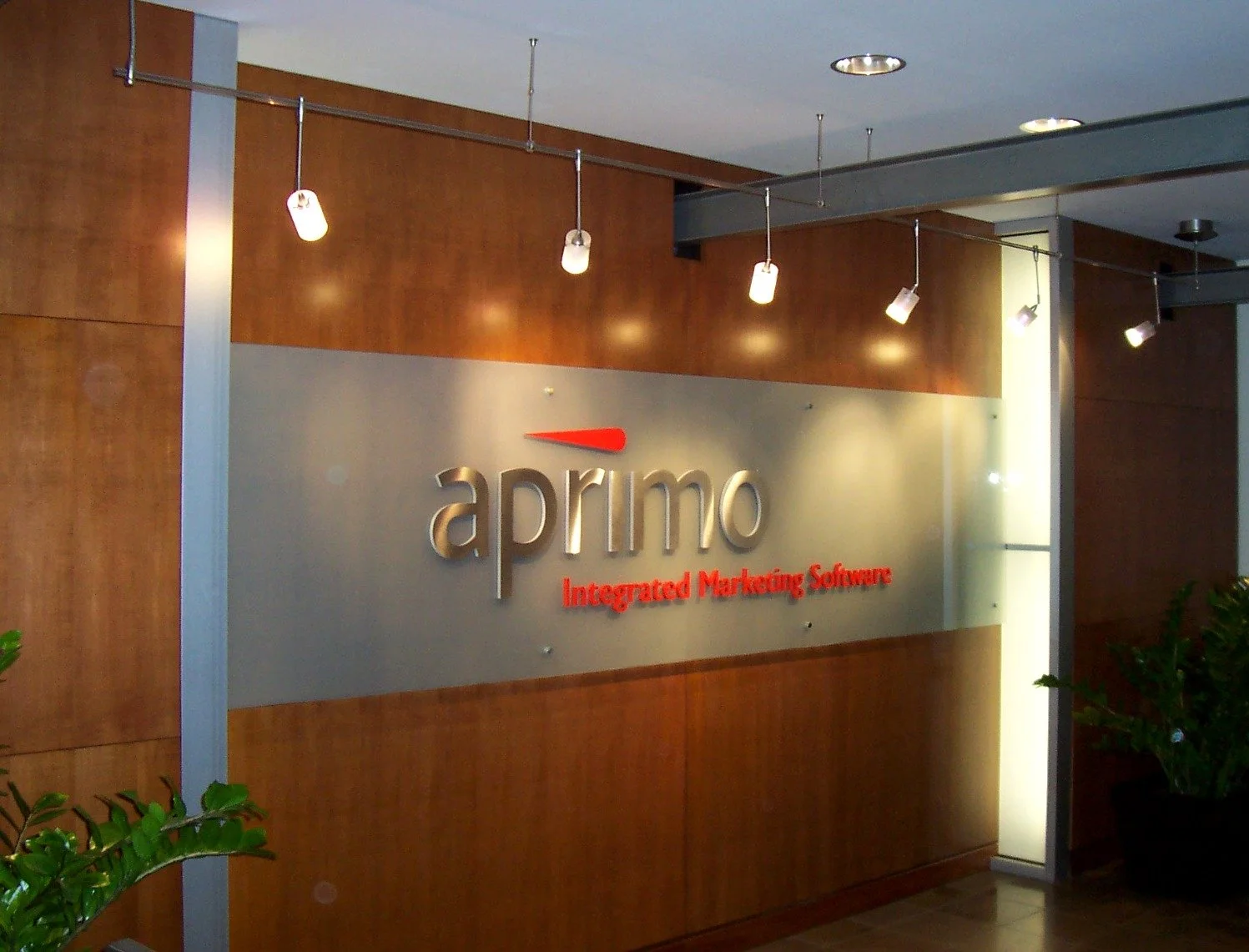 Office lobby wall with a sign displaying 'aprico' and the tagline 'Integrated Marketing Software' in red text, wooden paneling, and ceiling lights.