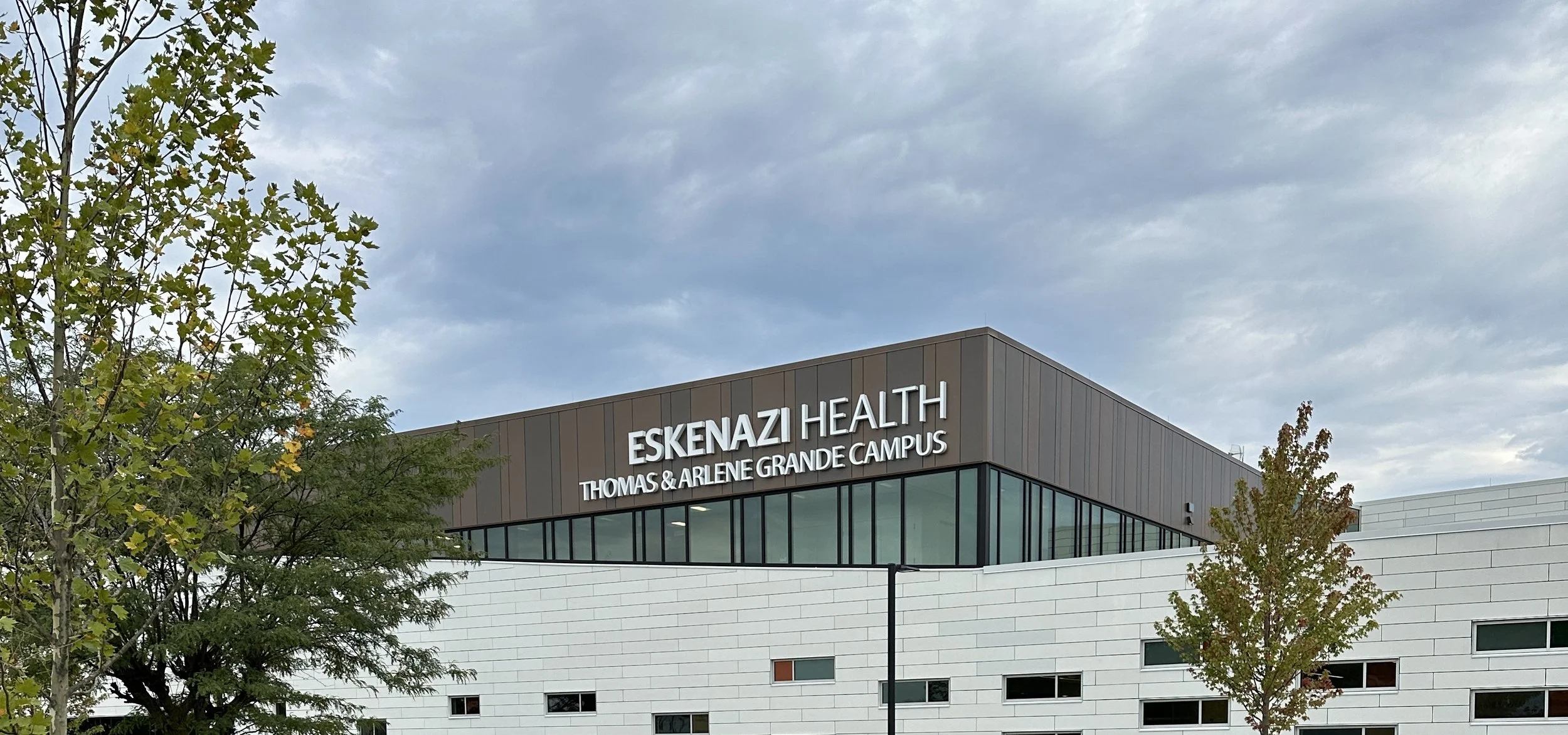 Exterior view of Eskenazi Health Thomas & Arlene Grande Campus building with trees in the foreground and a cloudy sky above.