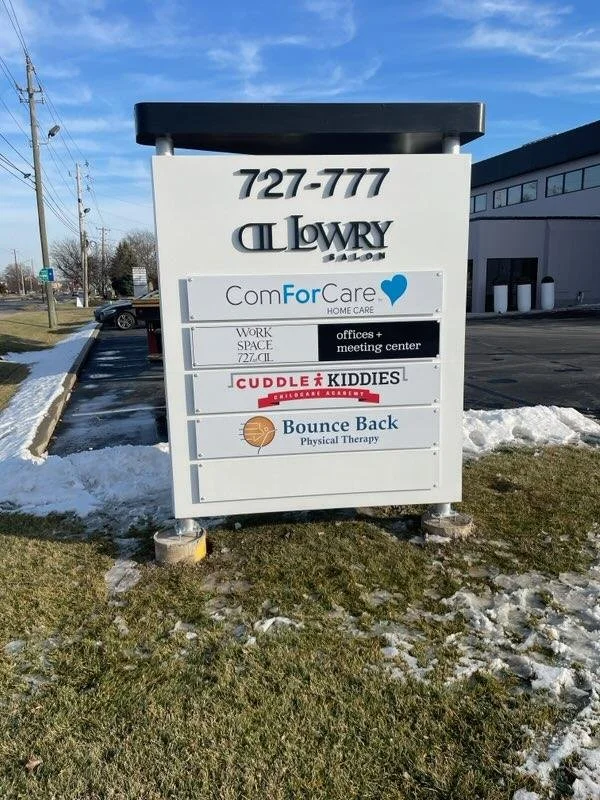 Signboard for a business complex with multiple tenants, including ComForCare Home Care, a work space with offices and meeting center, Cuddle Kiddies preschool, and Bounce Back physical therapy at 727-777 on a partly snow-covered lawn.