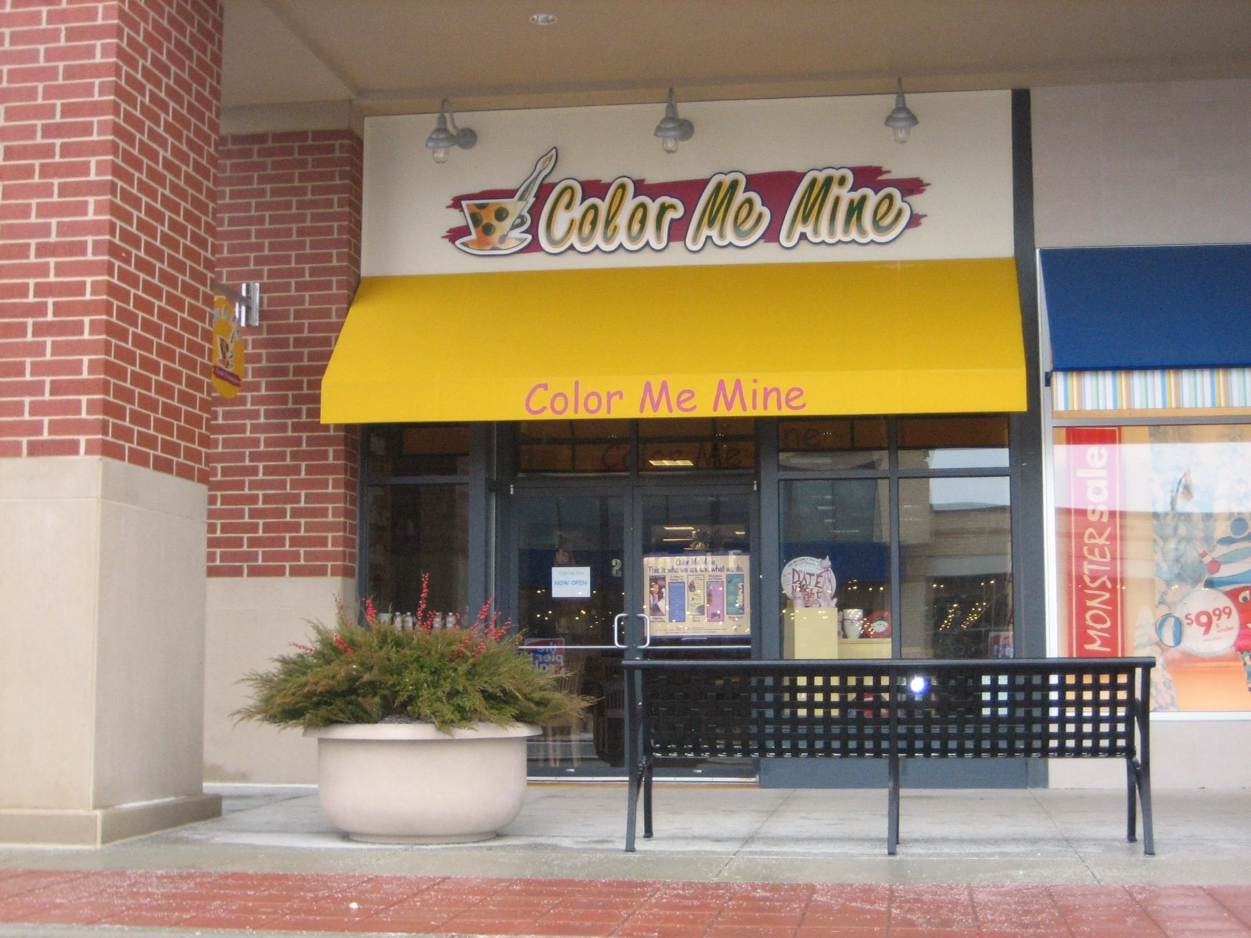 Storefront for Color Me Mine, a ceramic painting studio, with a yellow awning displaying the store's name. There is a large flower pot with greenery and a black bench outside the entrance, with a glass door and windows showing the interior.