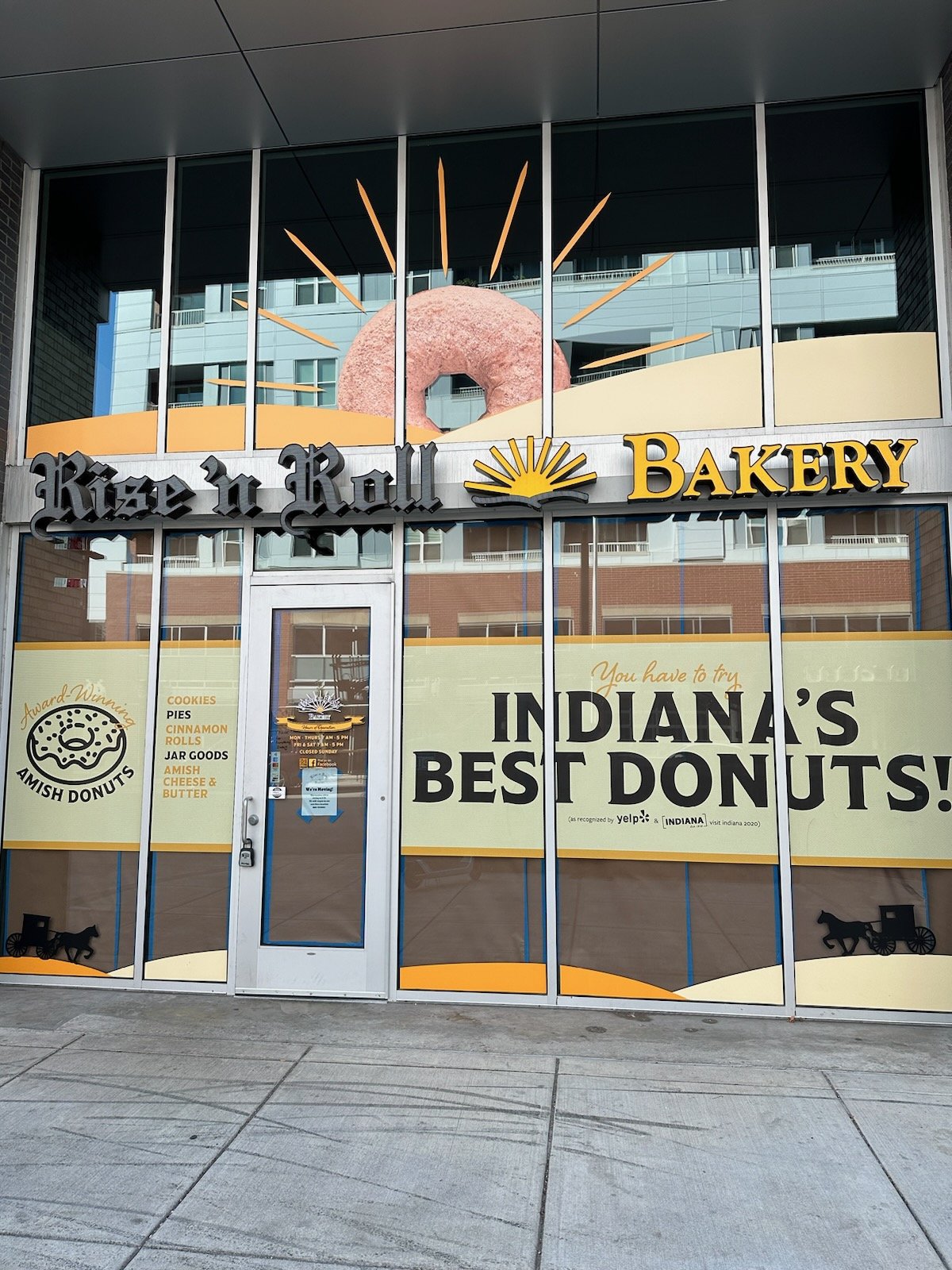 Storefront for Rise n Roll Bakery with large window display featuring a pink donut with sprinkles and a sunburst design, with signs advertising Amish donuts, cookies, pies, cinnamon rolls, jar goods, and cheese and butter, and a large banner stating 