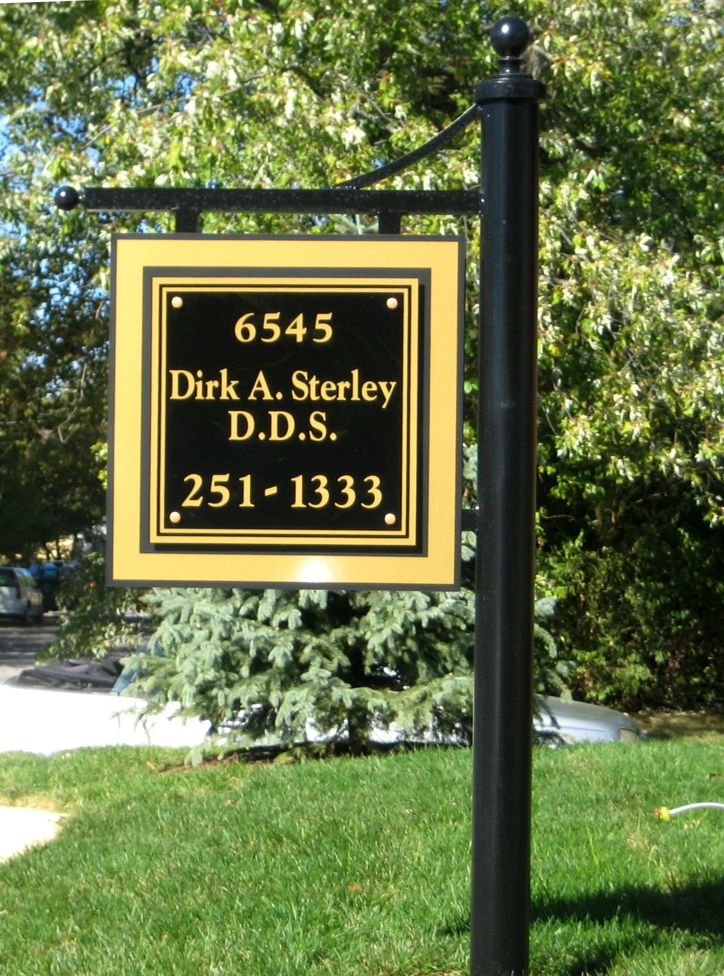 A black and yellow sign on a black metal pole with the address 6545, the name Dirk A. Sterley D.D.S., and the phone number 251-1333, set against a background of green trees and grass.