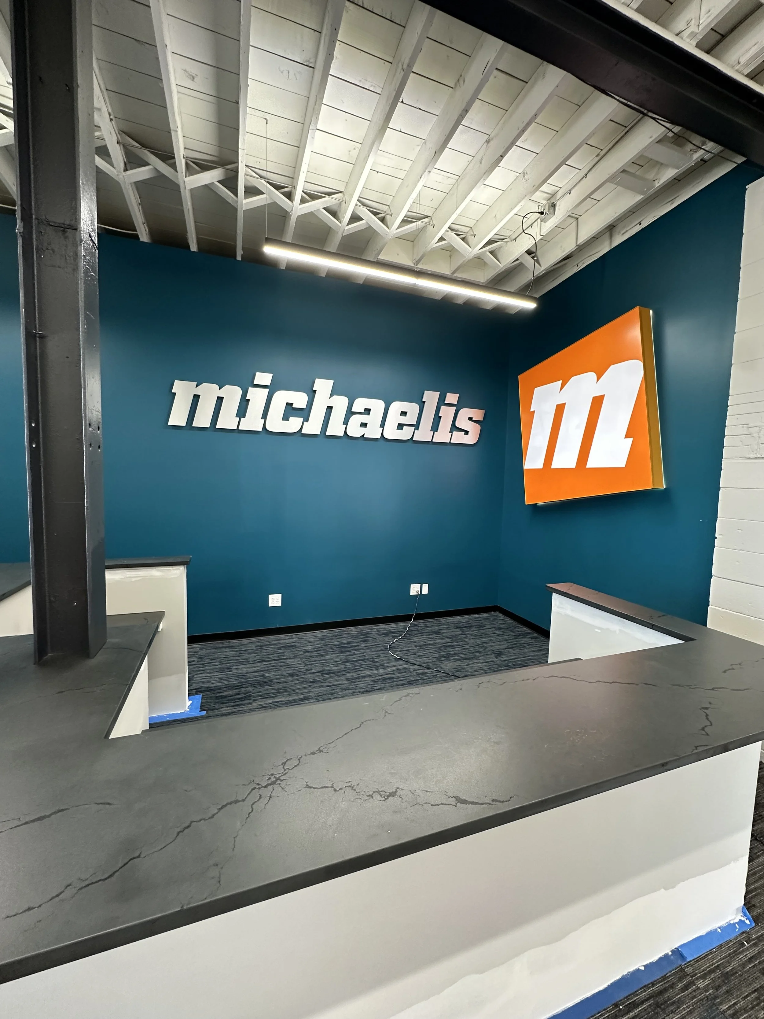 Interior of a store or office with a teal wall displaying the 'michaels' sign and the Michaels logo. The ceiling has exposed white beams and a linear light fixture. There is a black countertop in the foreground and some finished electrical outlets on