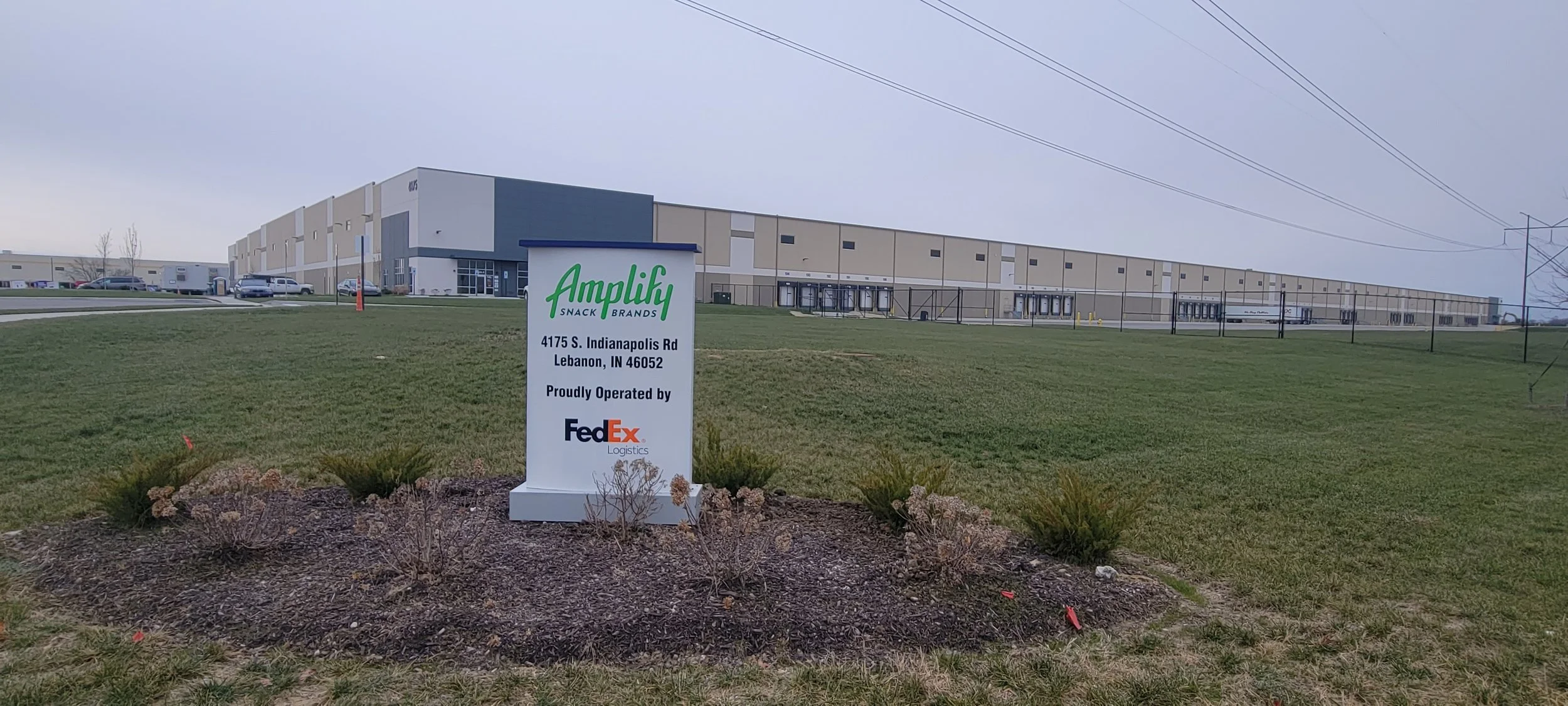 A large industrial warehouse building with multiple loading docks and parked trucks on a cloudy day, with a sign in the foreground that reads "Amplify Snack Brands, 4175 S. Indianapolis Rd, Lebanon, IN 46052, Proudly Operated by FedEx Logistics."