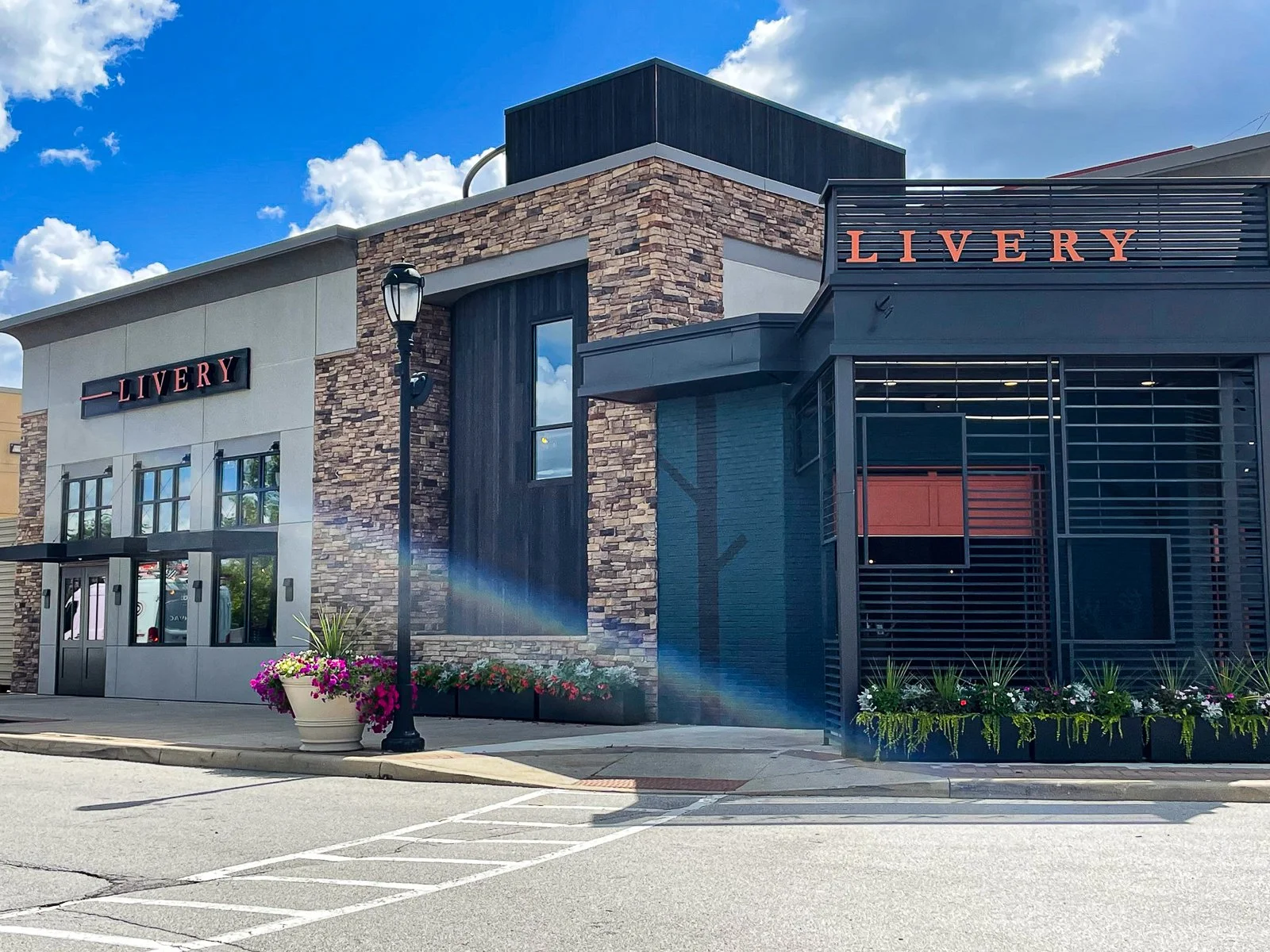 Exterior view of a modern retail building with signage reading 'LIVERY', featuring a mix of stone, dark wood, and metal elements, with potted flowers and a lamppost in the foreground under a partly cloudy sky.