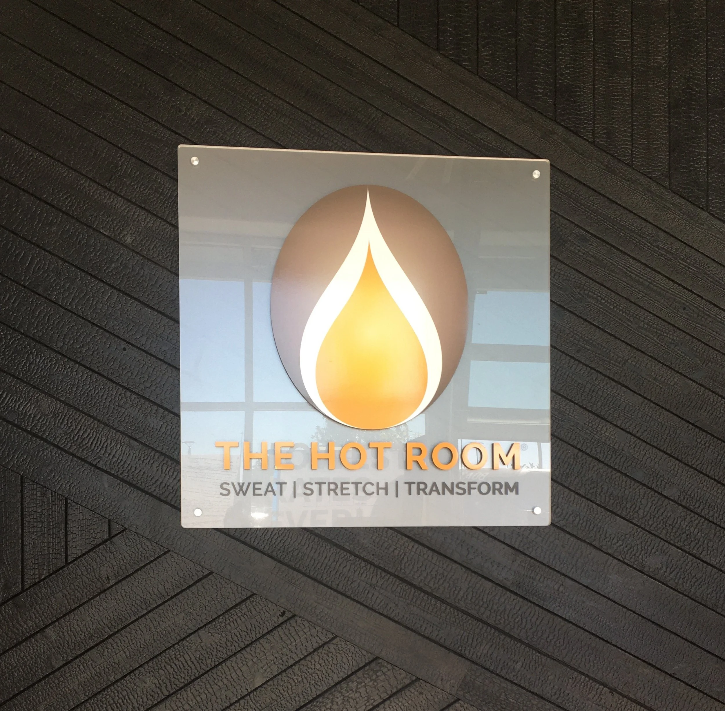 Sign for The Hot Room, a wellness center with a flame logo, mentioning services like sweat, stretch, and transform, mounted on a dark textured wall.