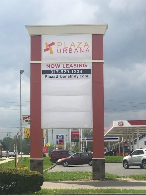 Large vacant freestanding sign for Plaza Urbana with leasing information, located in a shopping area with cars and a gas station in the background.