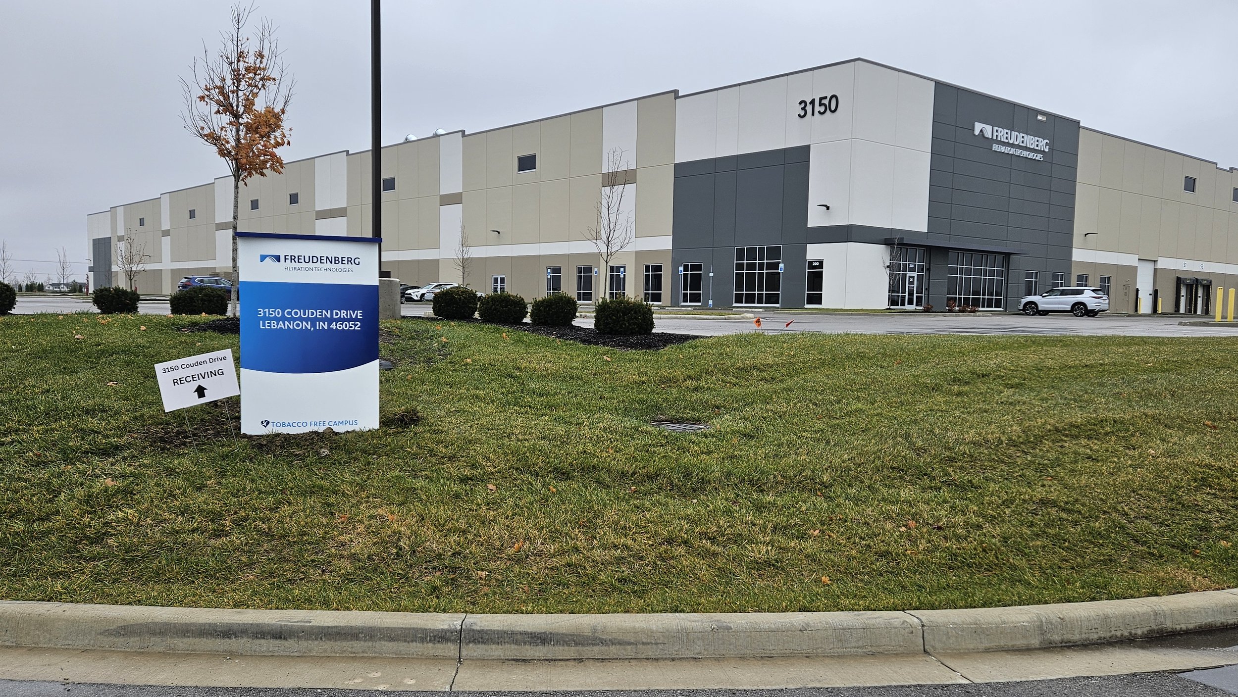 Exterior of a modern industrial building with a sign indicating the address 3150 Couden Drive, Lebanon, Indiana. The building has gray and white paneling, multiple entrances, and a parking lot with several cars. The grass in the foreground has small 