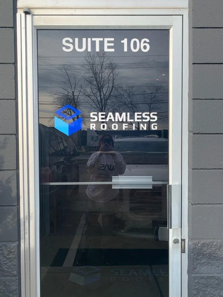 Glass door with a sign that reads 'SUITE 106' at the top and the logo and name of 'SEAMLESS ROOFING' in the center. The reflection in the glass shows a person taking the photo, some vehicles, and trees.