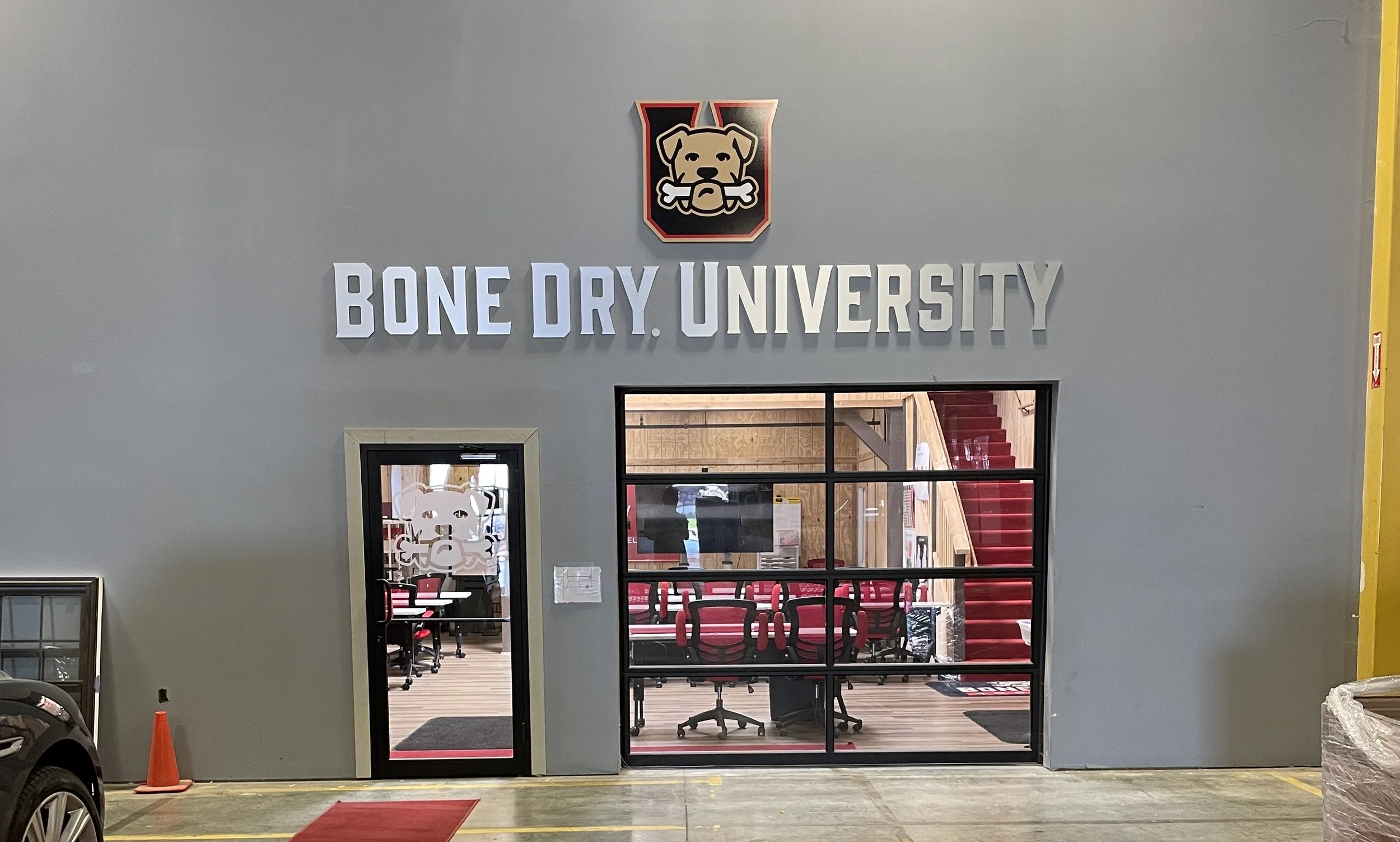 Entrance to Bone Dry University, showing a gray wall with a sign and logo, a door, and a window revealing a classroom with chairs and stairs inside.