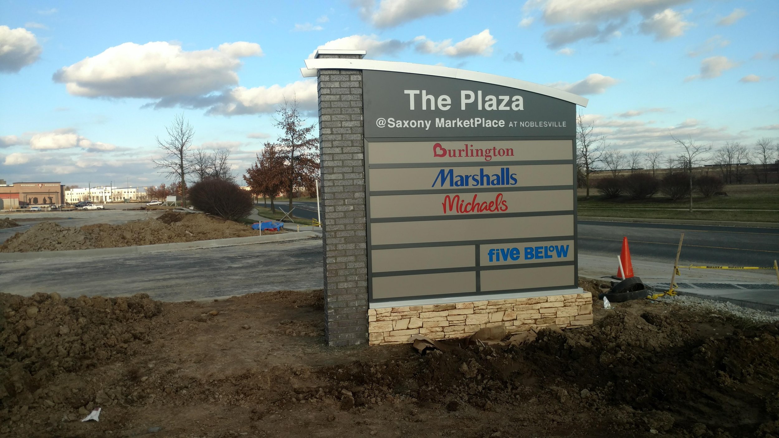 Sign for The Plaza at Saxony MarketPlace in Noblesville listing stores Burlington, Marshalls, Michaels, and Five Below, with construction in progress around it.