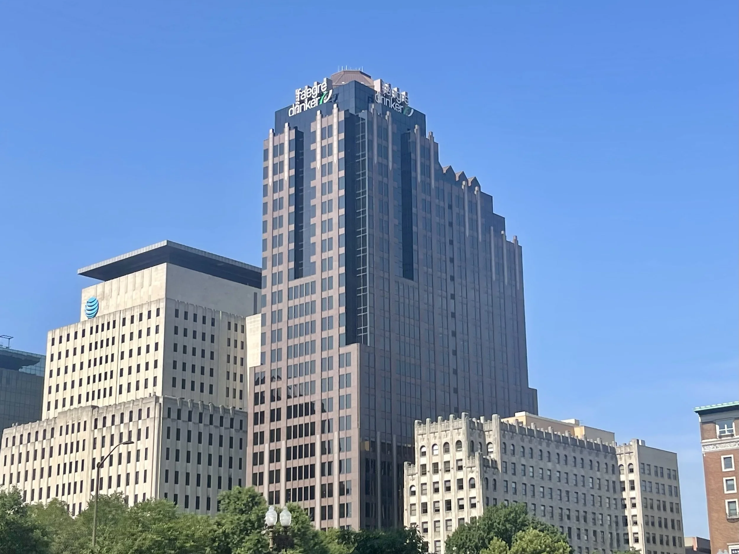 Tall modern skyscraper with a sign at the top reading 'Fajre dranken' against a clear blue sky, surrounded by other buildings and greenery at the base.