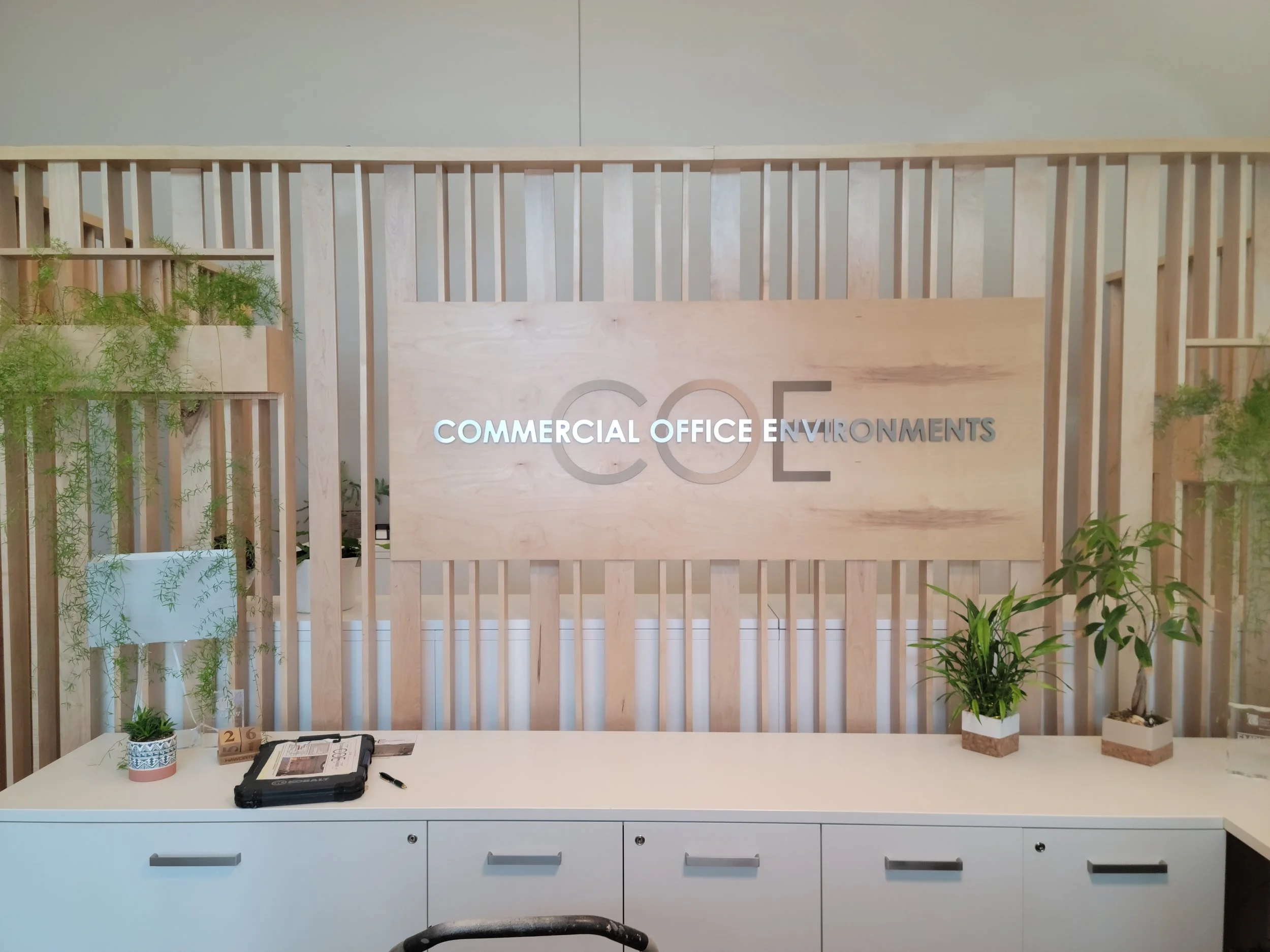 Reception area with a clerical desk, potted plants, and a wooden privacy screen featuring a sign that reads 'Commercial Office Environments' in an office setting.