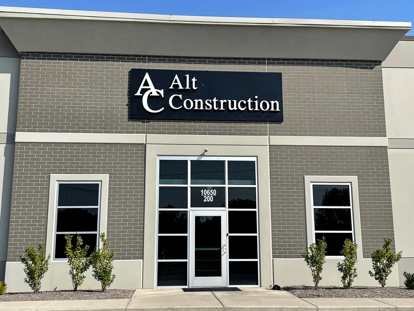 Exterior of a commercial building with a sign reading 'Alt Construction,' with glass doors and windows, small bushes in front, and a brick and light-colored wall facade.