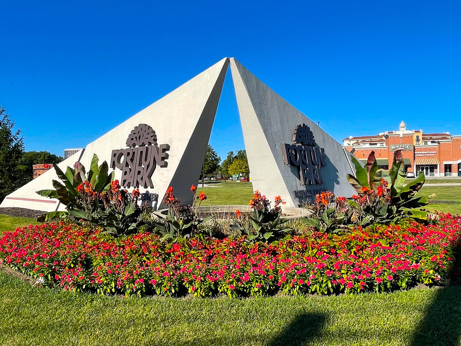 Entrance sign for Fortune Park with a flower bed in front and a building with a McLuster's sign in the background under a clear blue sky.