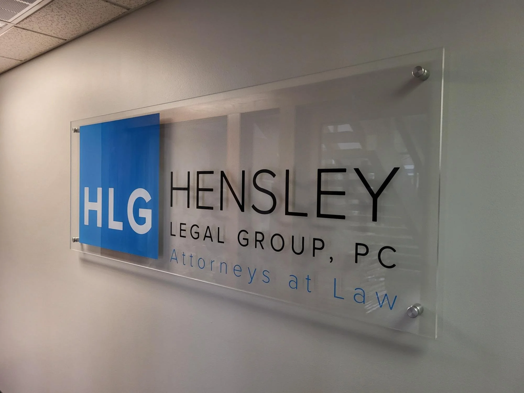 A glass sign on a wall for Hensley Legal Group, P.C., attorneys at law, with a blue rectangle containing white letters 'HLG' and black text for the firm's name and services.
