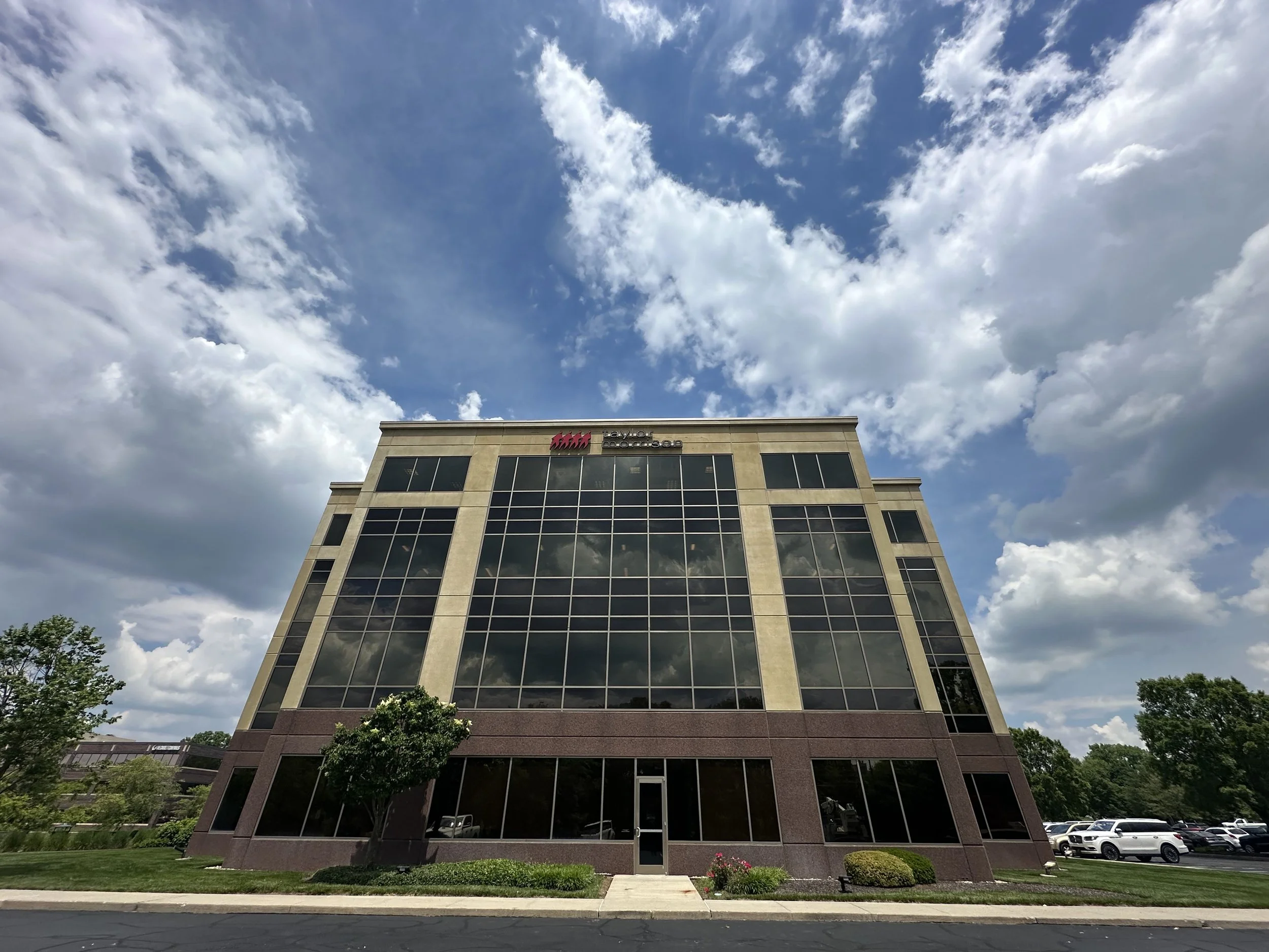 A modern office building with glass windows and a beige facade, set against a partly cloudy sky. There are trees and parked cars in the surrounding area.