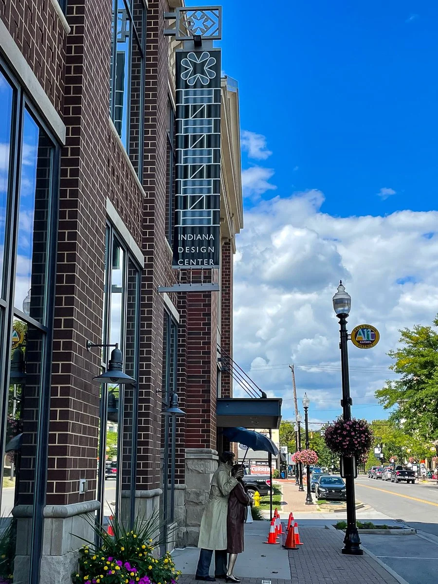 Street view with people under umbrellas, brick building with Indiana Design Center sign, street lamps, flower baskets, and traffic cones.