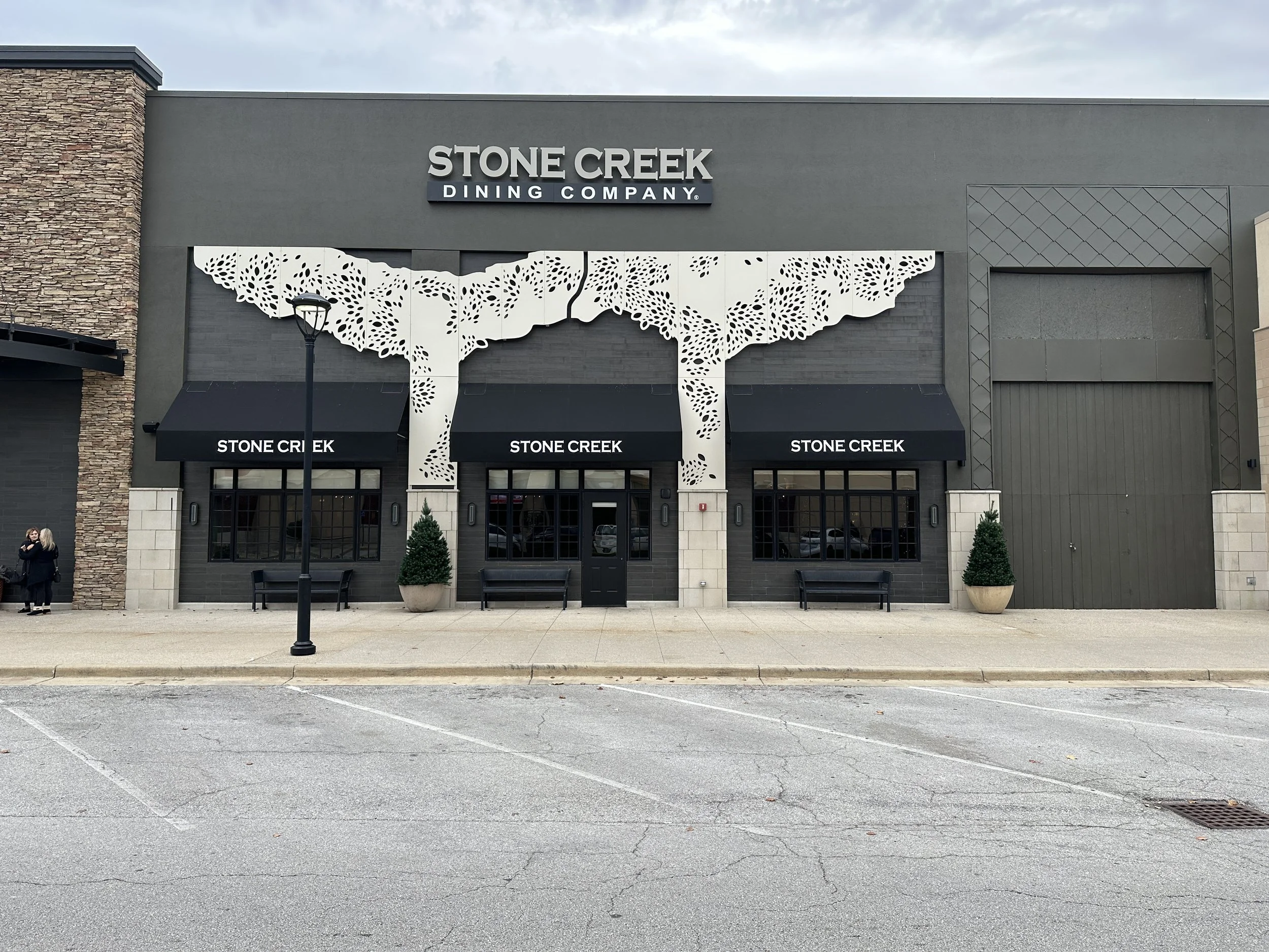 Storefront with black awnings reading "Stone Creek" and a decorative white metal tree sculpture attached above, on a sidewalk in front of a parking lot.