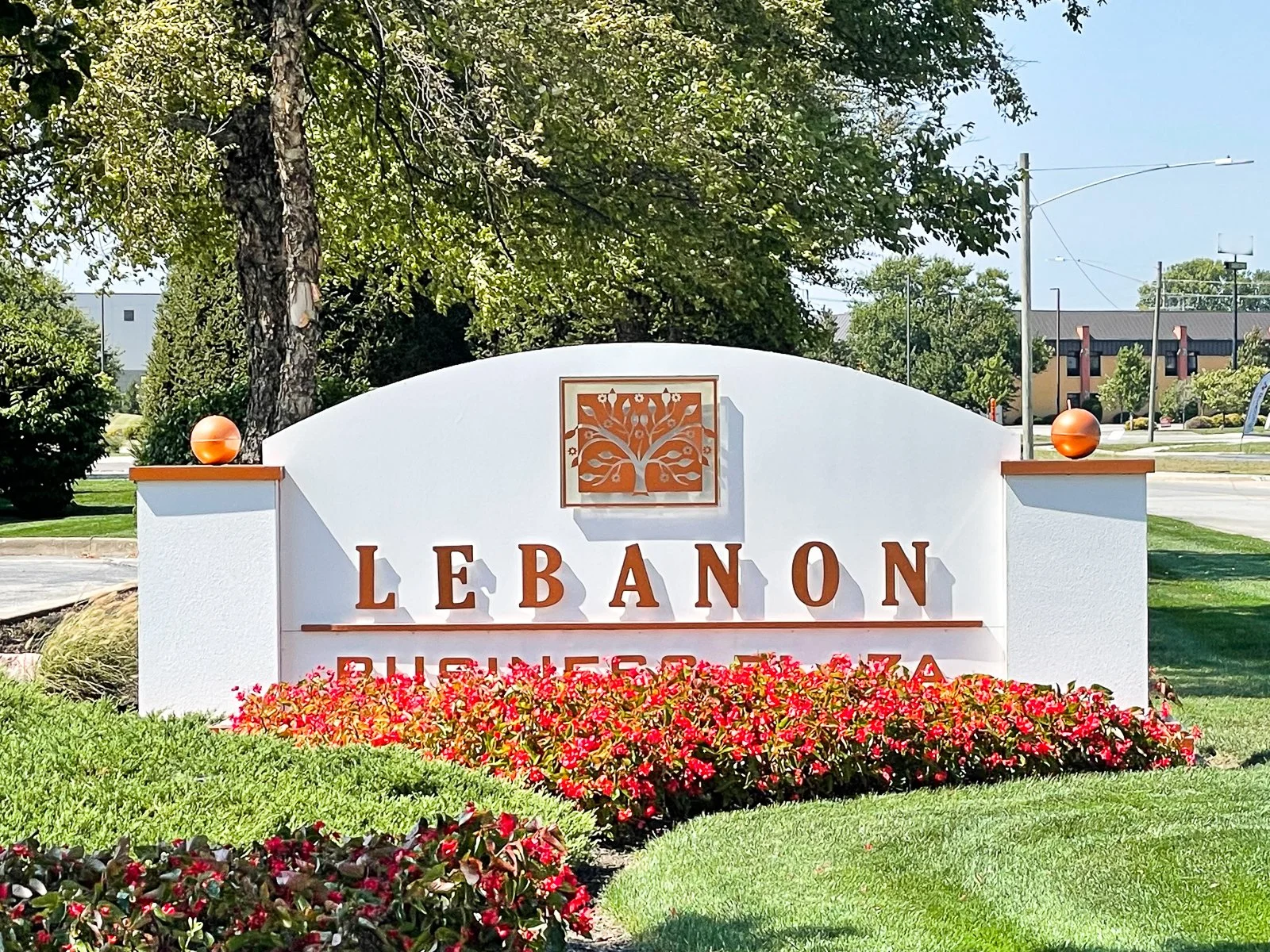 Sign reading "Lebanon Business Park" with a decorative tree logo above, surrounded by red and pink flowers, green grass, and trees in the background.