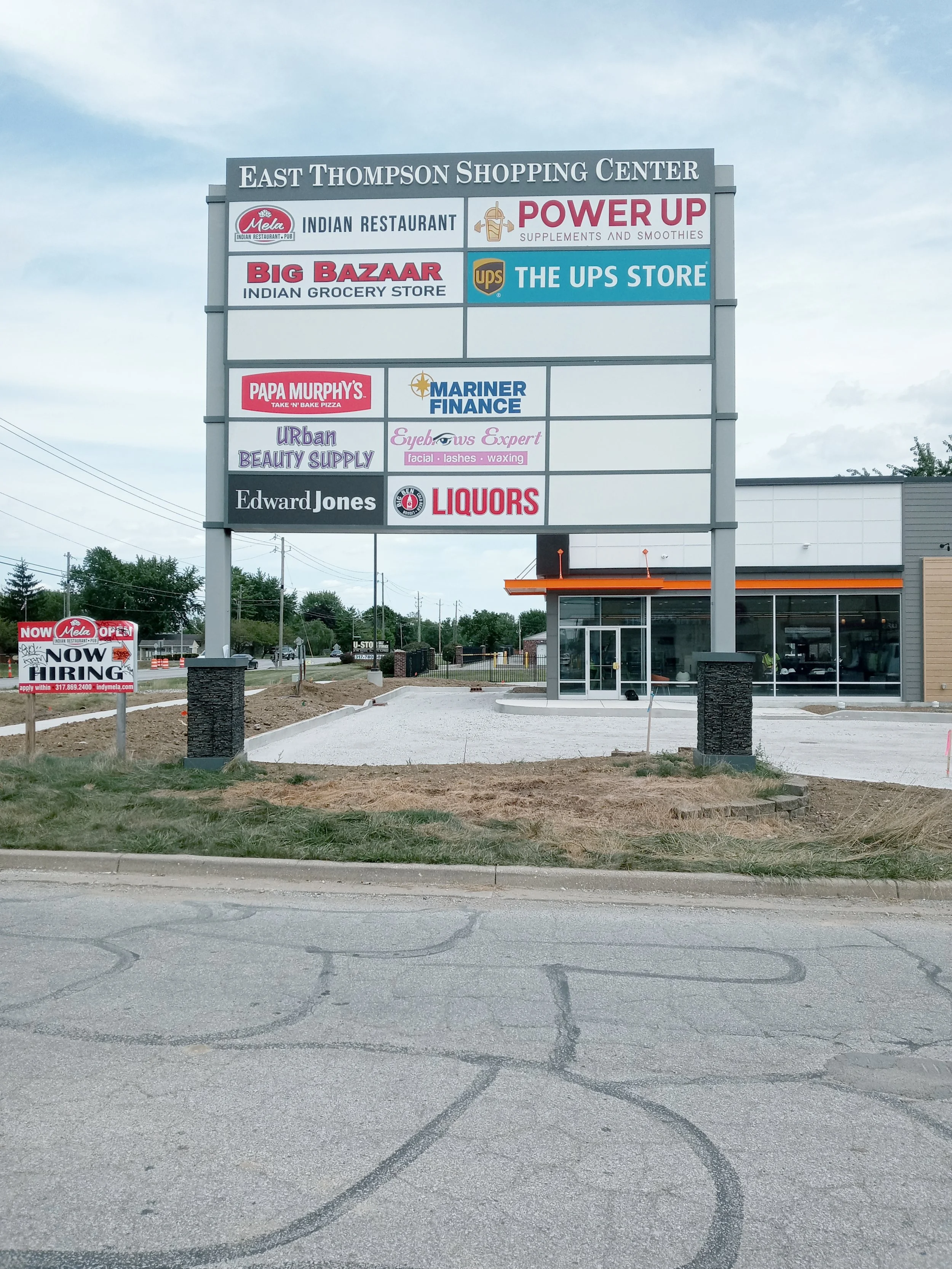 A large outdoor shopping center sign for East Thompson Shopping Center displaying various store names including Indian restaurant, supplements and smoothies, Indian grocery store, UPS Store, pizza restaurant, finance, beauty supply, eyelash and waxin