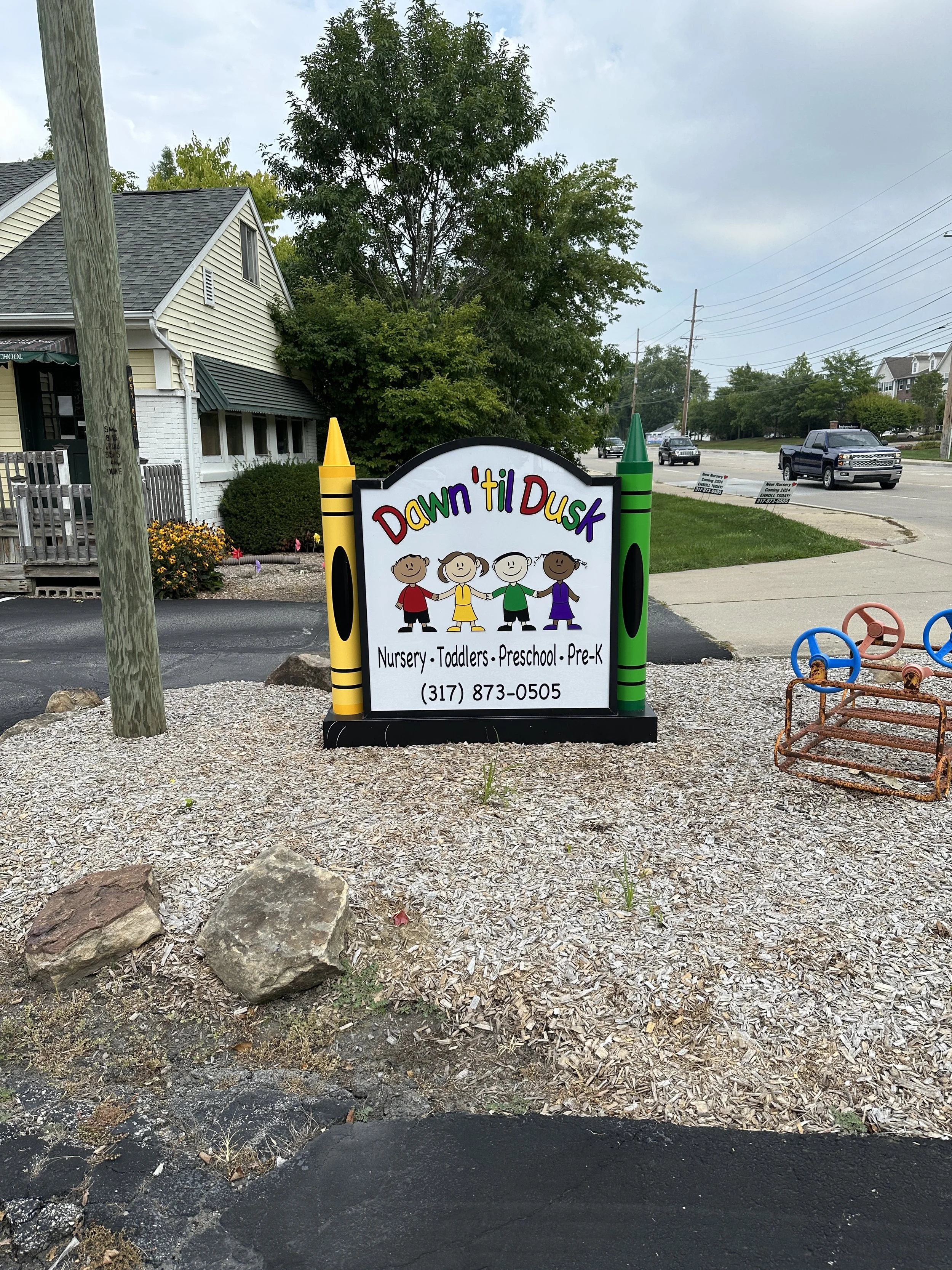 Sign for Dawn 'til Dusk childcare center with colorful text, cartoon children holding hands, and contact information, placed in a yard with rocks and mulch.
