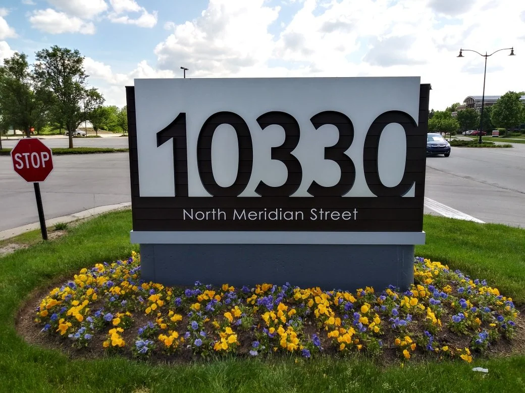 A sign displaying the address 10330 North Meridian Street, surrounded by yellow and purple flowers, with a stop sign and cars visible in the background.