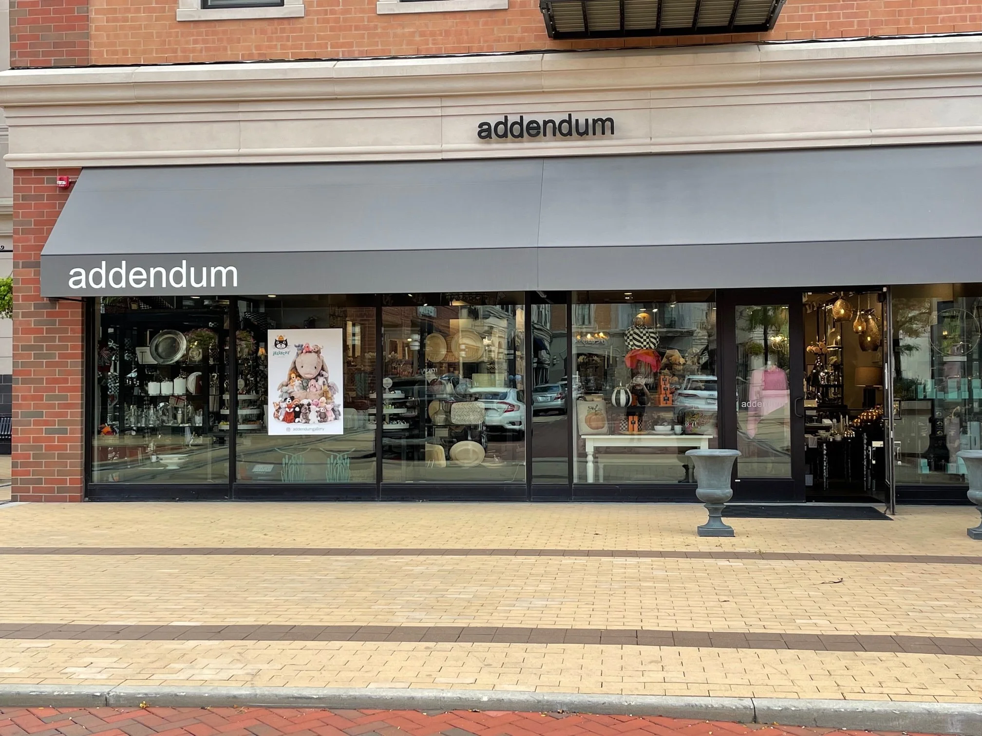 Storefront named 'addendum' with large glass windows displaying home decor items and a poster of a stuffed animal with accessories outside.