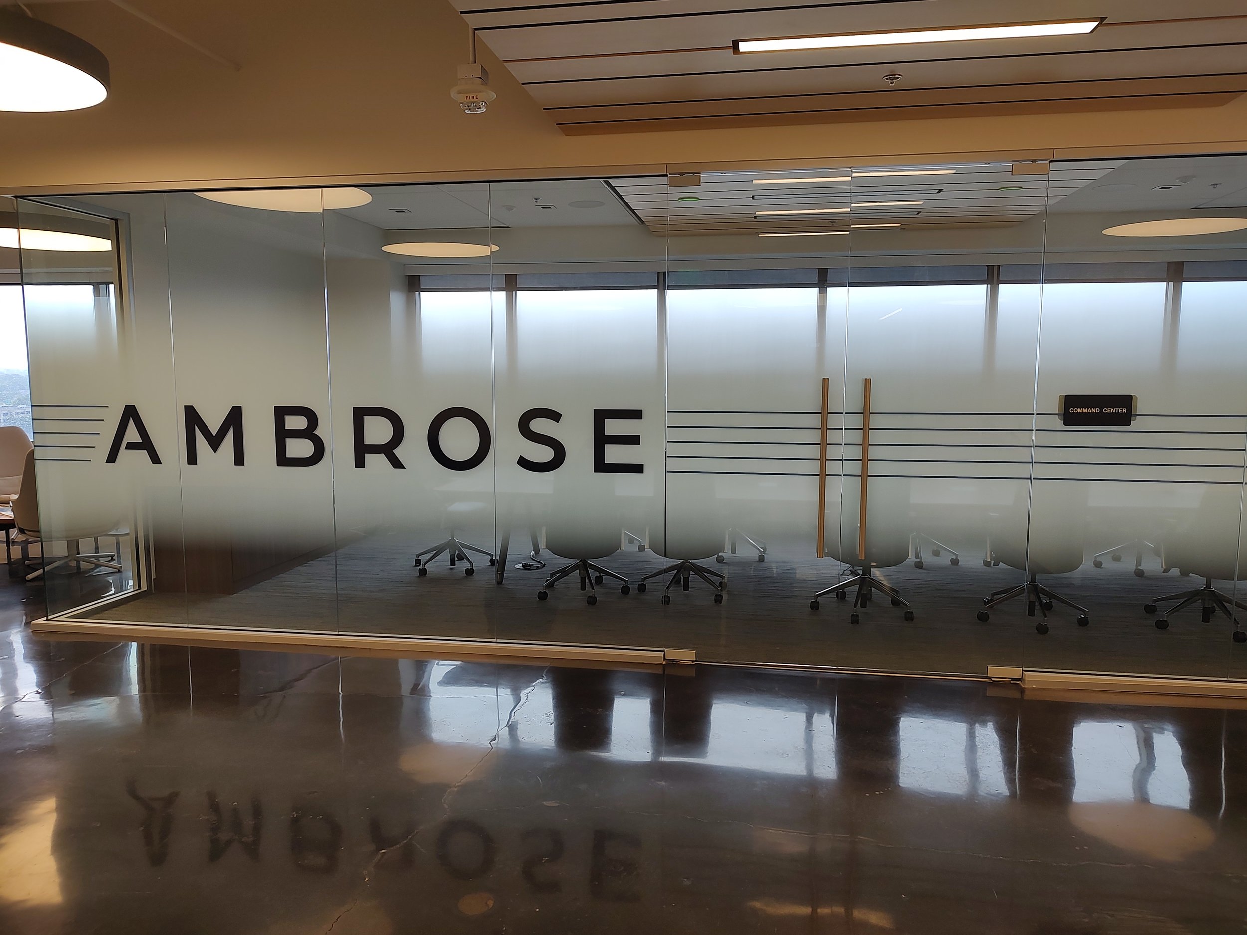 Office conference room with large glass wall displaying the word 'AMBROSE' and several white office chairs inside.
