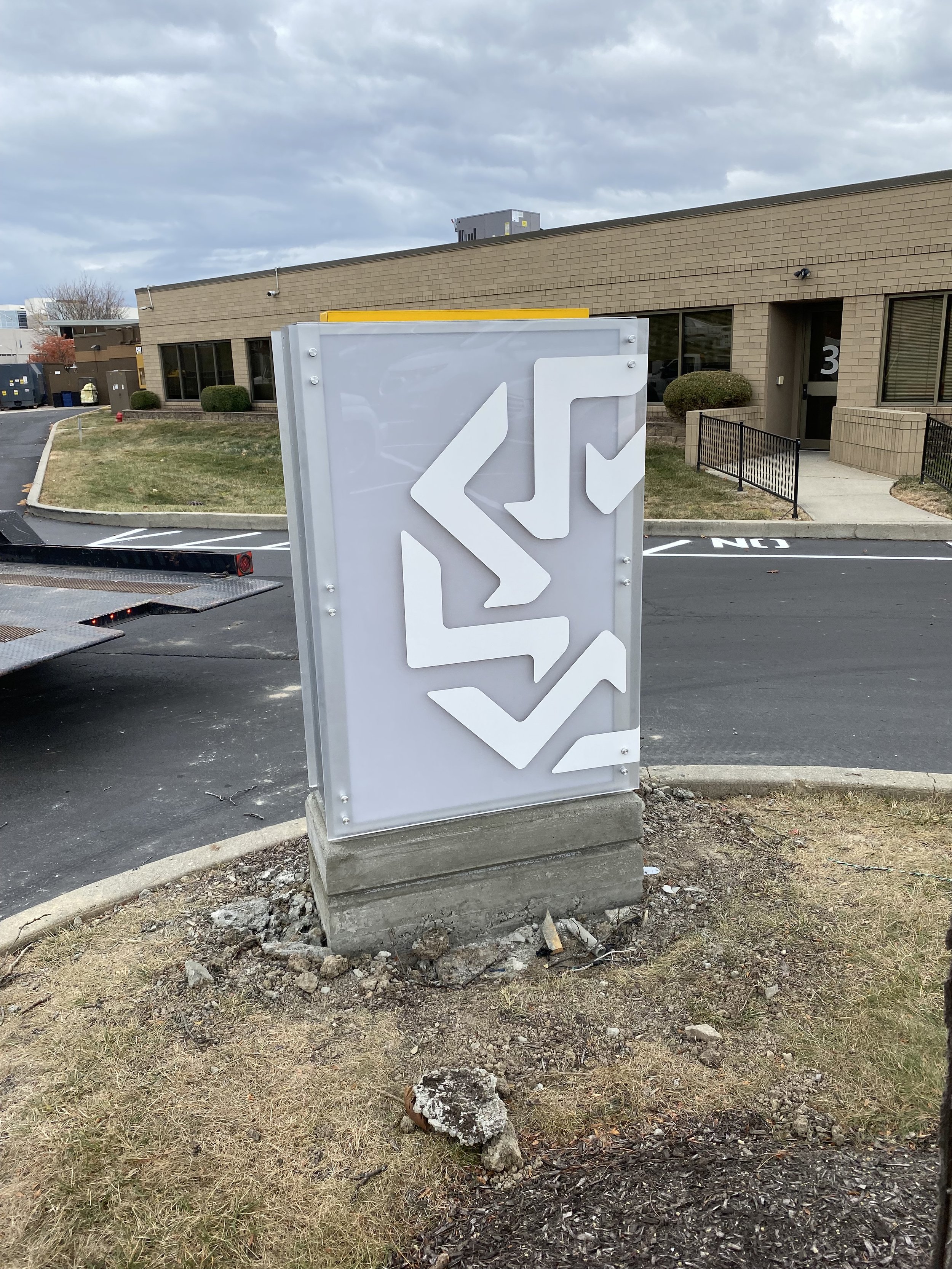 A large directional sign with arrows pointing left and right, located outside a commercial building with a road, grass, and shrubs in the background.