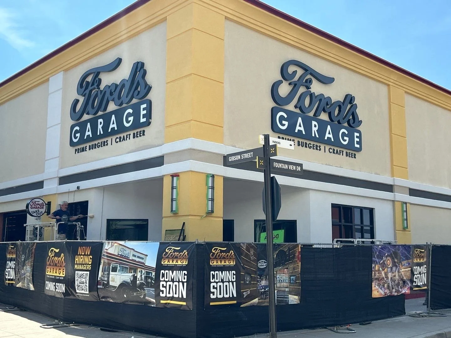 A building with large Ford's Garage signs and banners indicating the restaurant will open soon, located at the corner of Gibson Street and Fountain View Drive. The building is three stories high with a sidewalk and street signs visible in the foregro